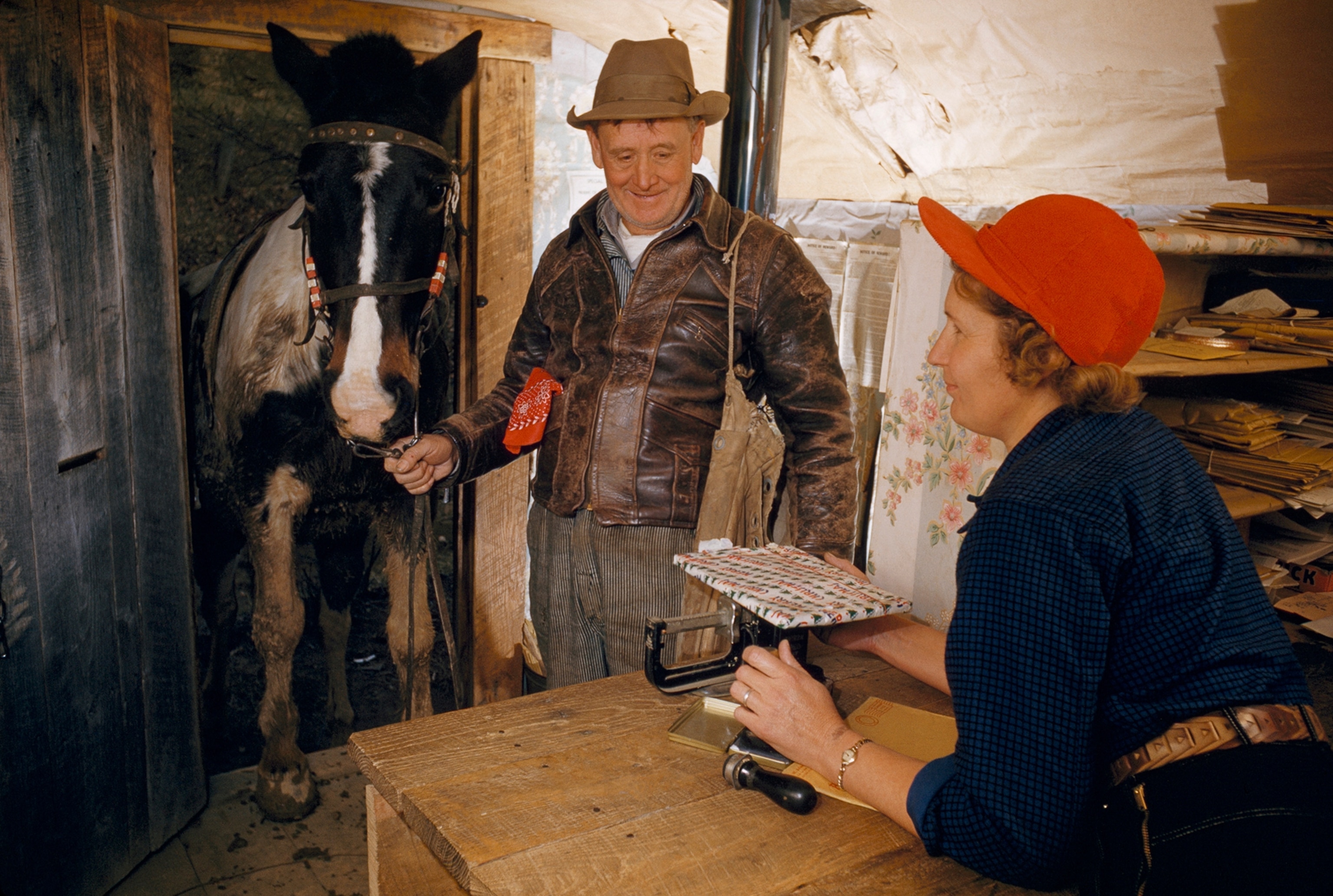 A postman and his horse wait for the postmistress to weigh a letter.