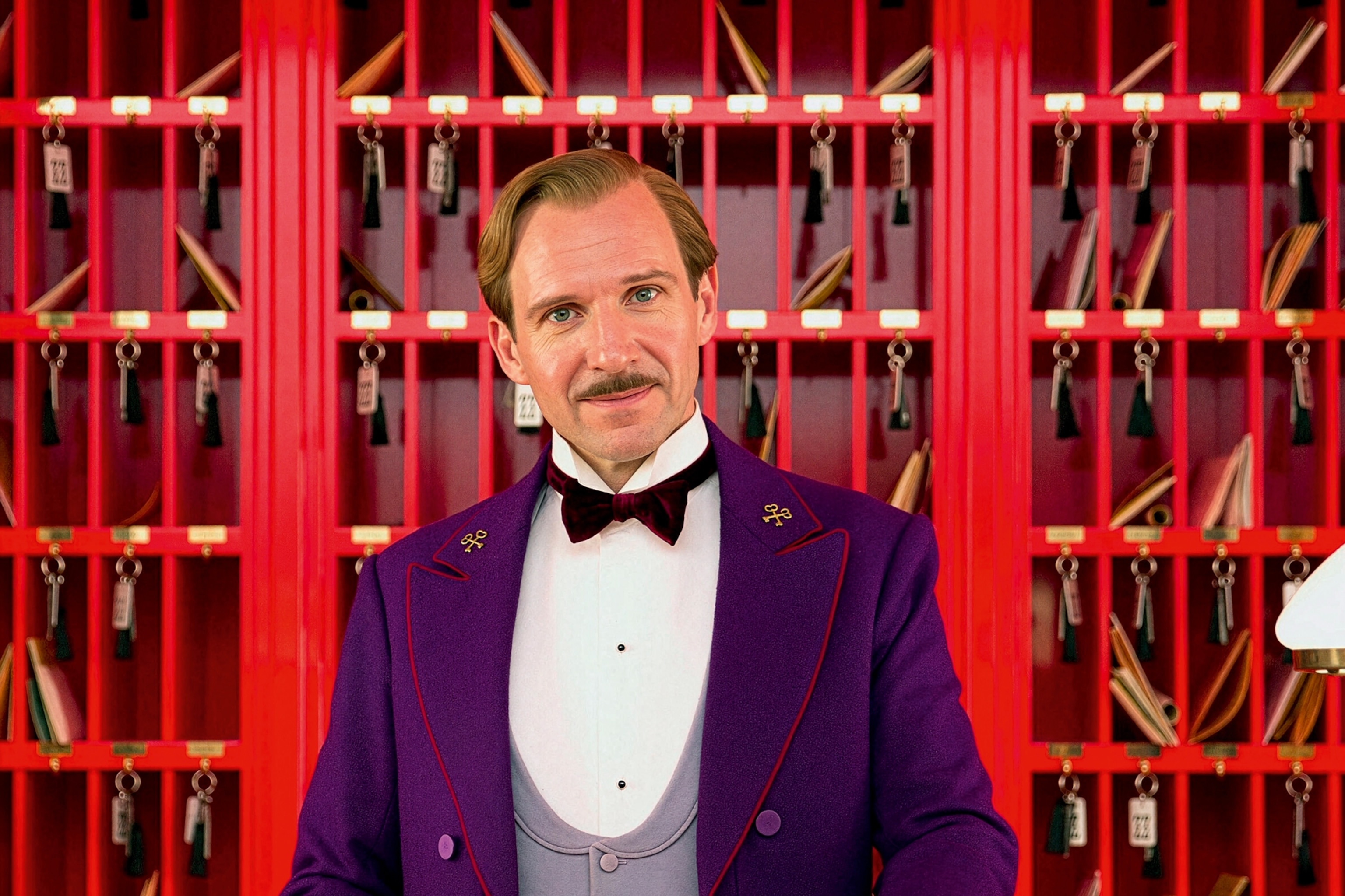 Ralph Fiennes in the role of concierge Gustave H from The Grand Budapest Hotel behind a reception counter.