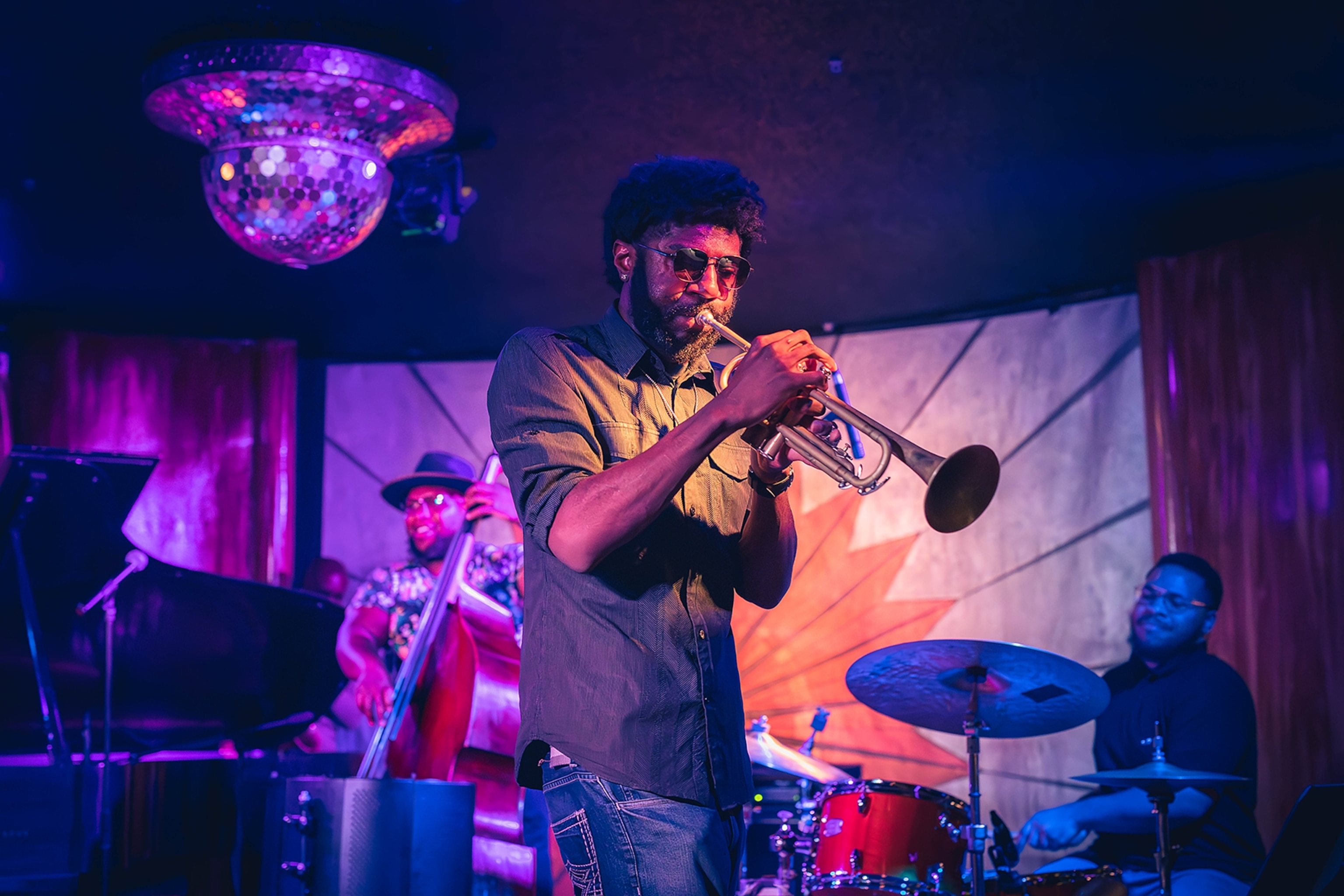 Man plays a trumpet illuminated by purple lighting with musicians playing a cello and the drums in the background