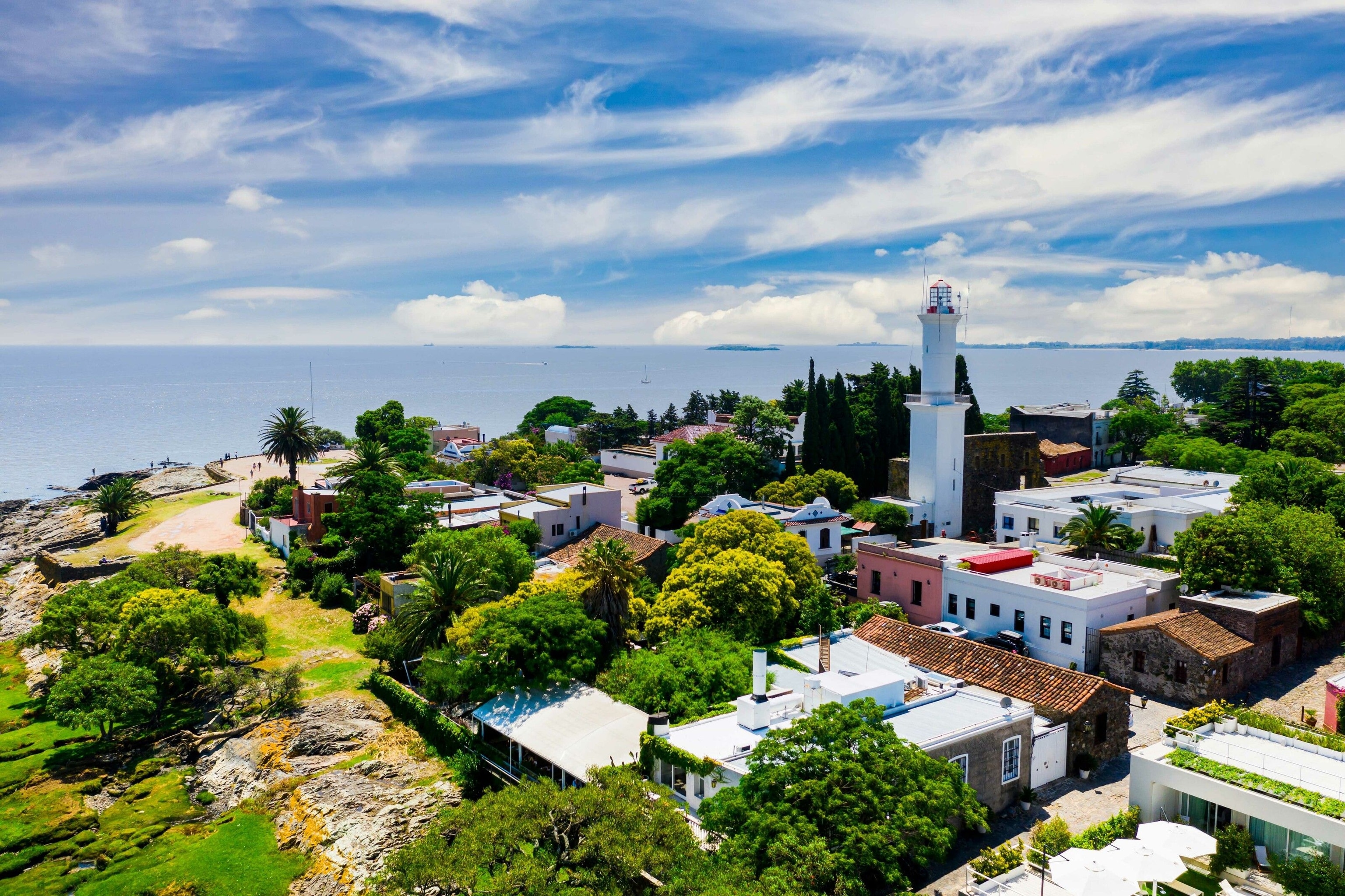 Colonia del Sacramento. The buildings are mostly white, including a tall white tower that looks like a lighthouse. Several small boats can be seen on the ocean. Islands sit in the distance.