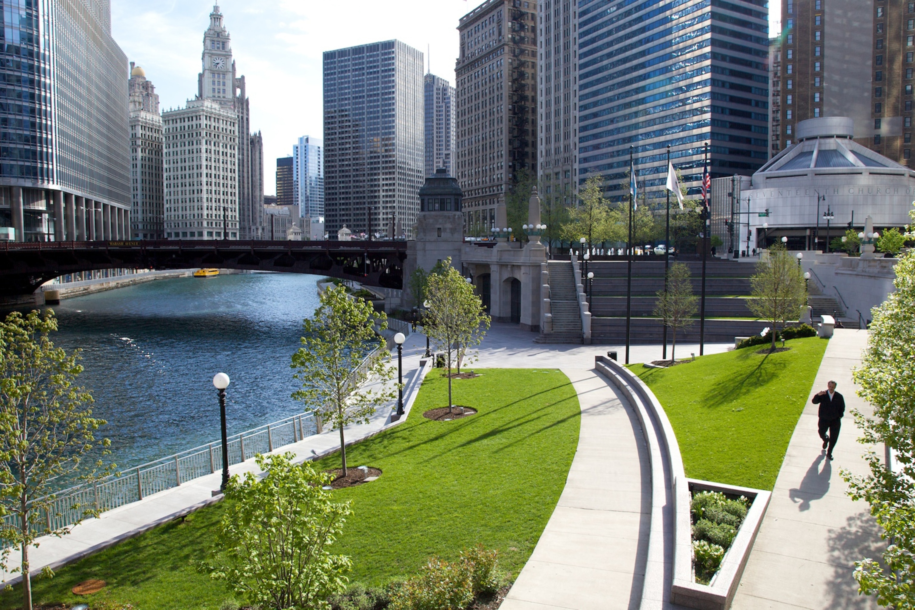An elevated view of a pedestrian walking along a paved and well manicured walkway beside the Chicago River.