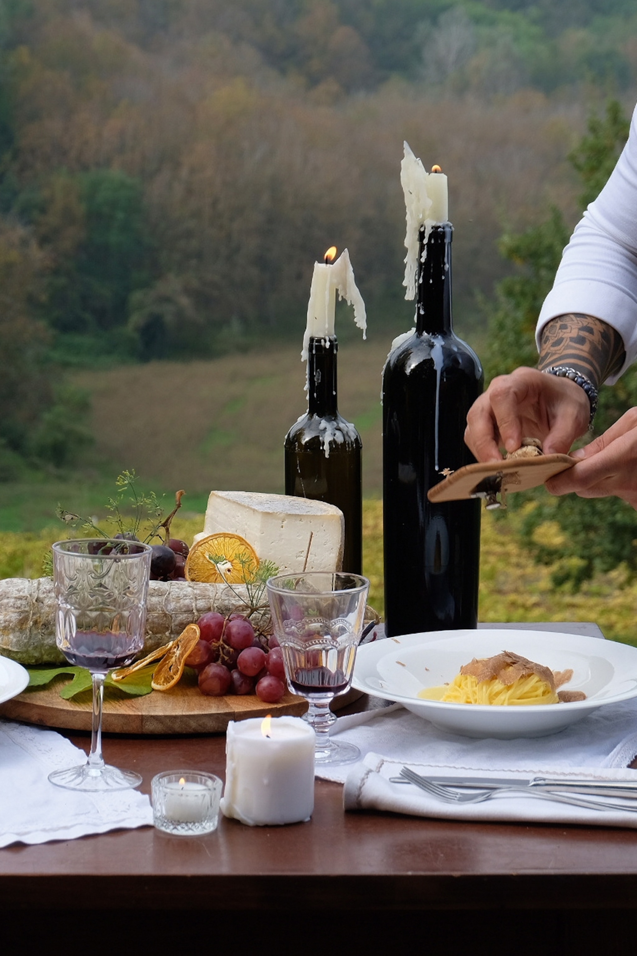 A waiter shaving truffles over a plate of pasta on a set table in a garden with wine and candles.