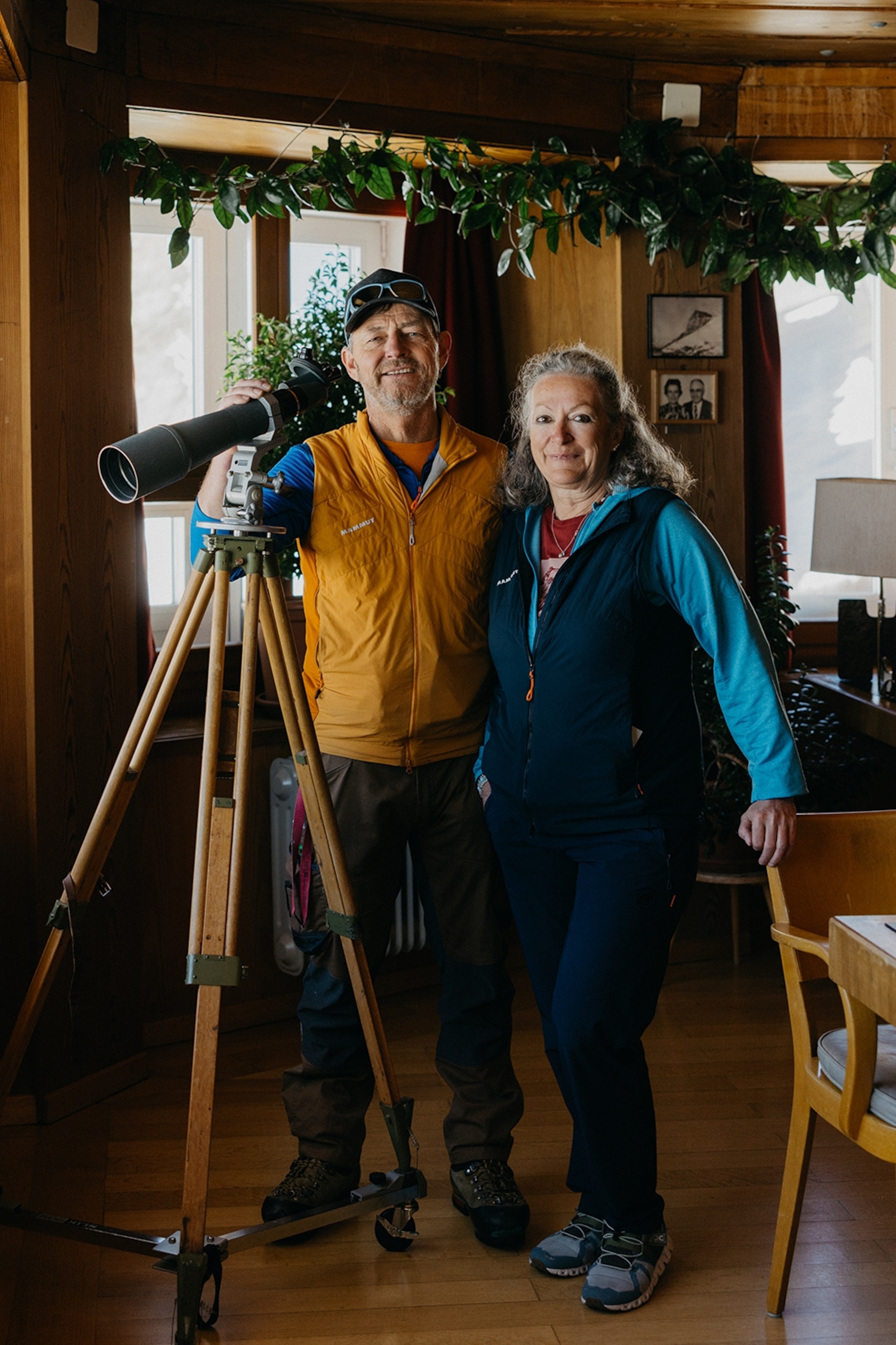 A middle-aged couple wearing hiking gear and posing for a full-body portrait in the wooden interiors of a high-altitude hut.