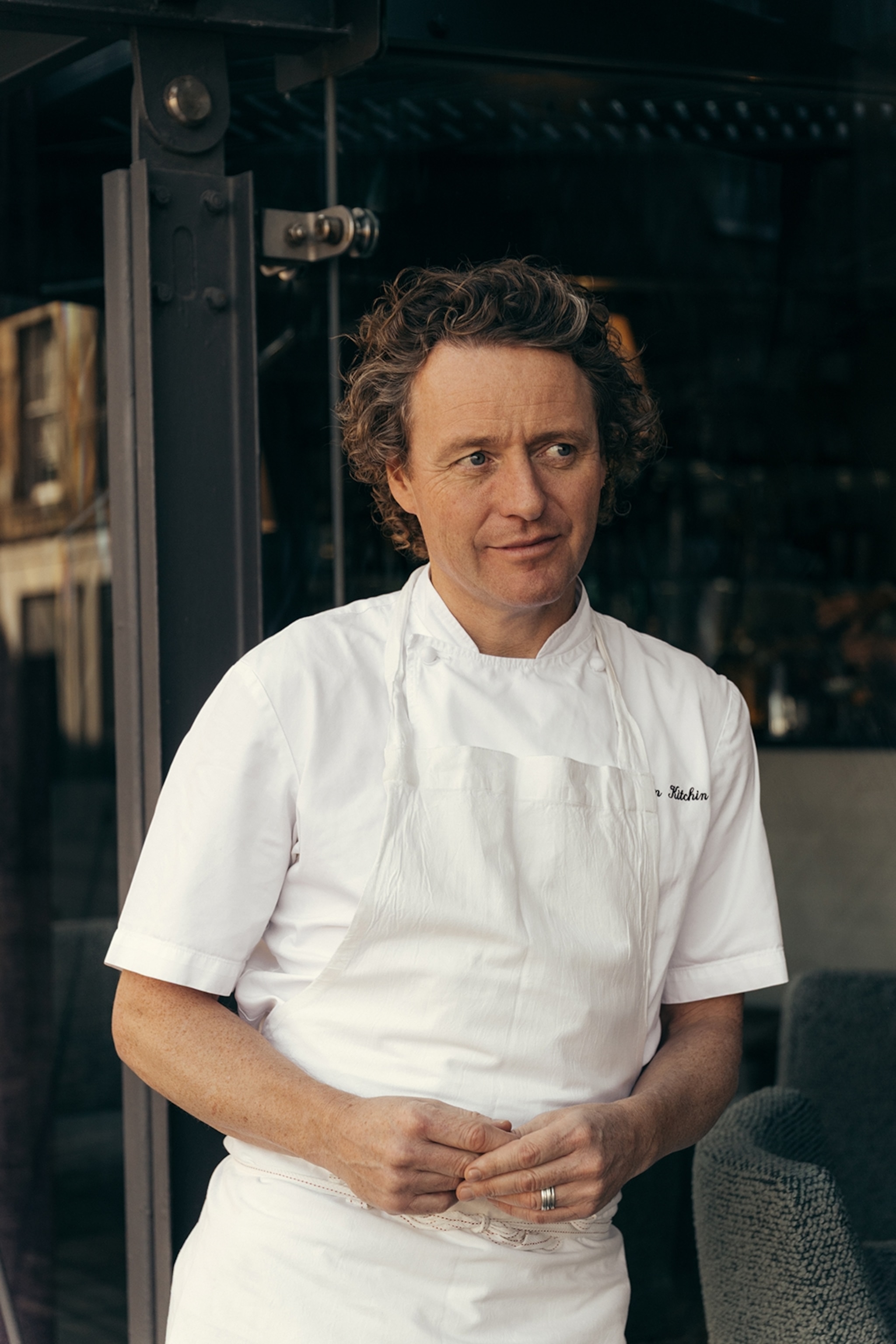 A middle-aged chef with wavy short hair leaning against a high chair in his chef coat and apron.