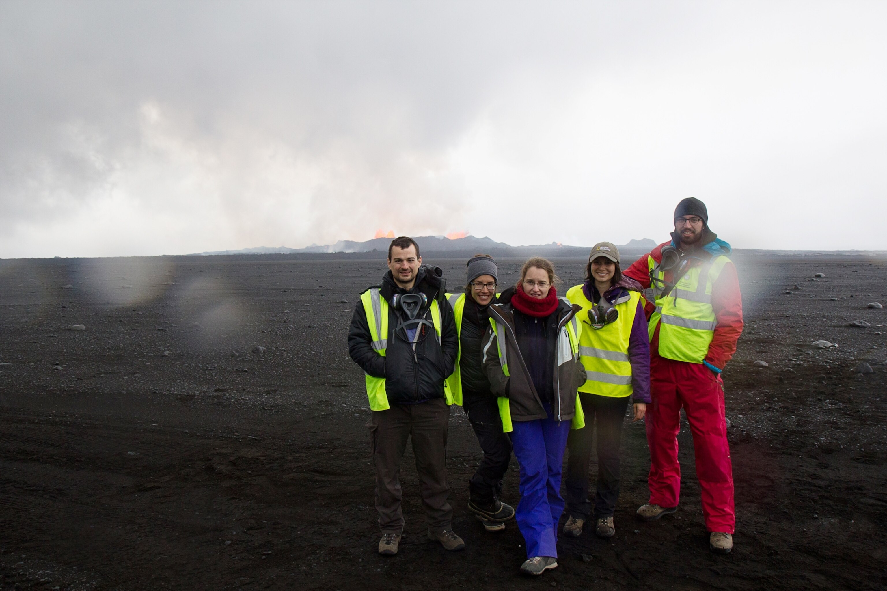 a group of scientists and a videographer standing together in front of the eruption at the Holuhraun fissure, red lava is erupting out of the site, the sky is smoky and gray