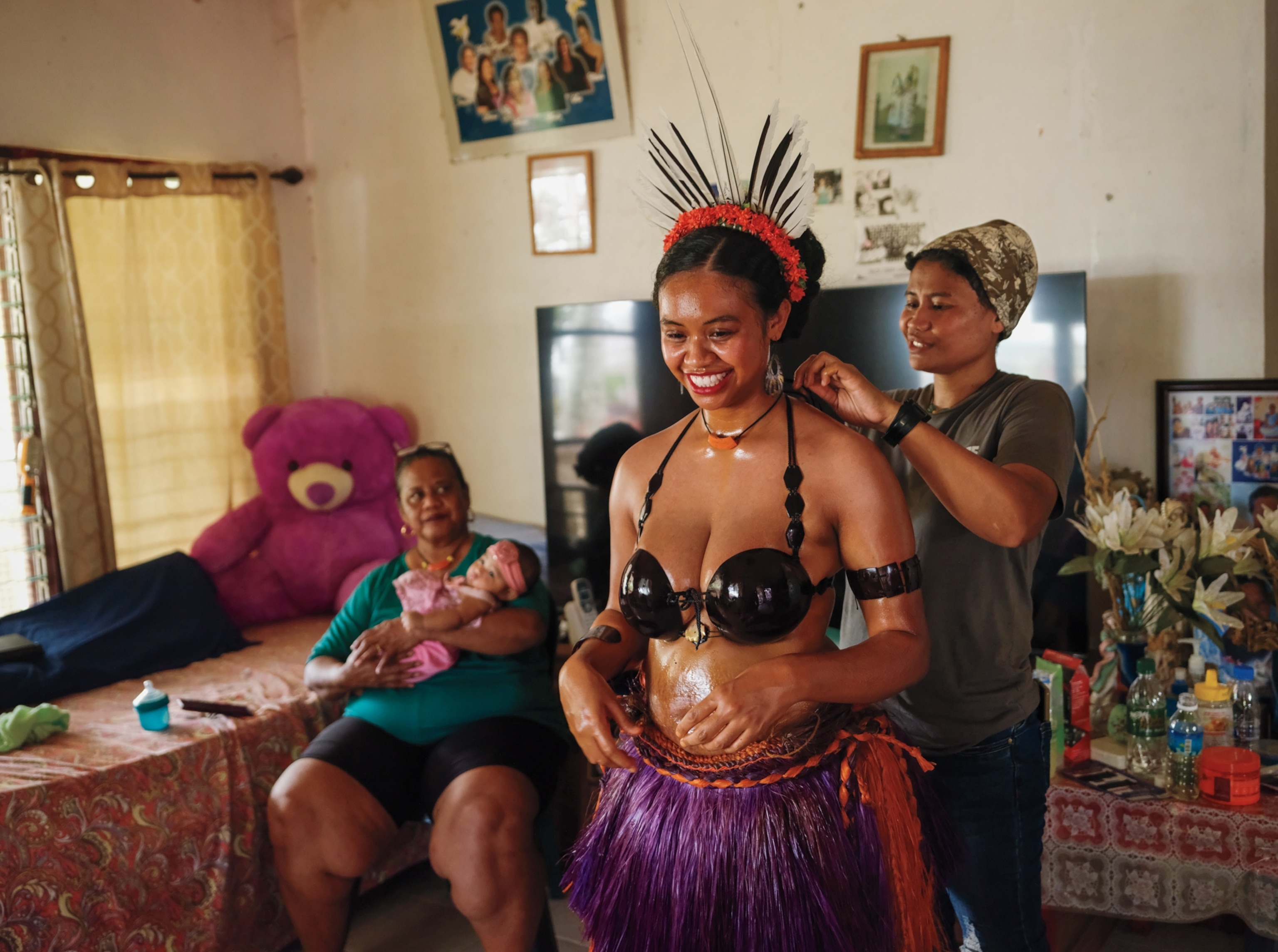 A woman wearing a grass skirt, small top, and headpiece stands as a woman behind her ties her top's strap behind her neck. In the background, another woman sits in a chair holding a new baby.