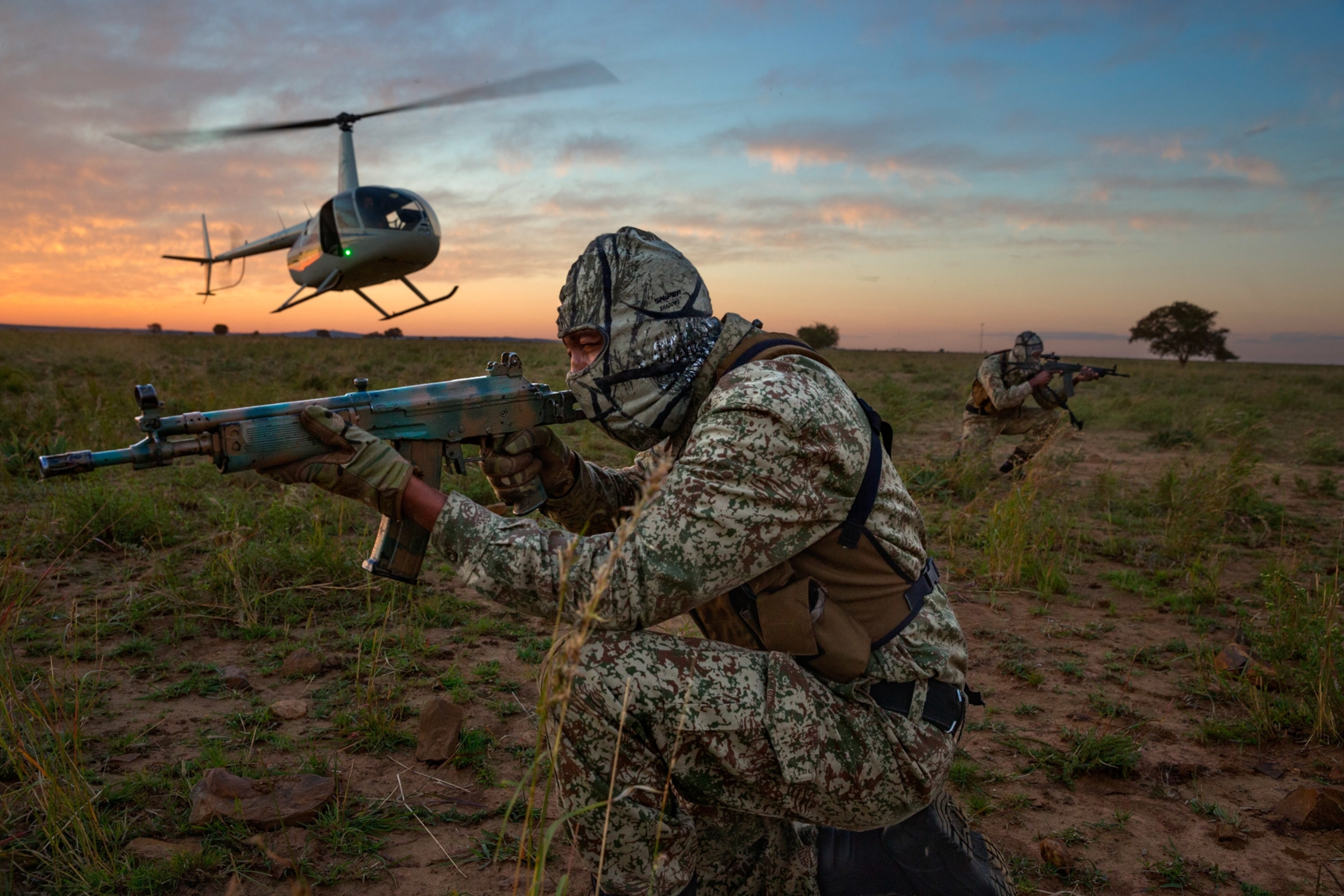 A team of two antipoachers are seen looking down the barrels of their riffles as a helicopter takes off behind them.