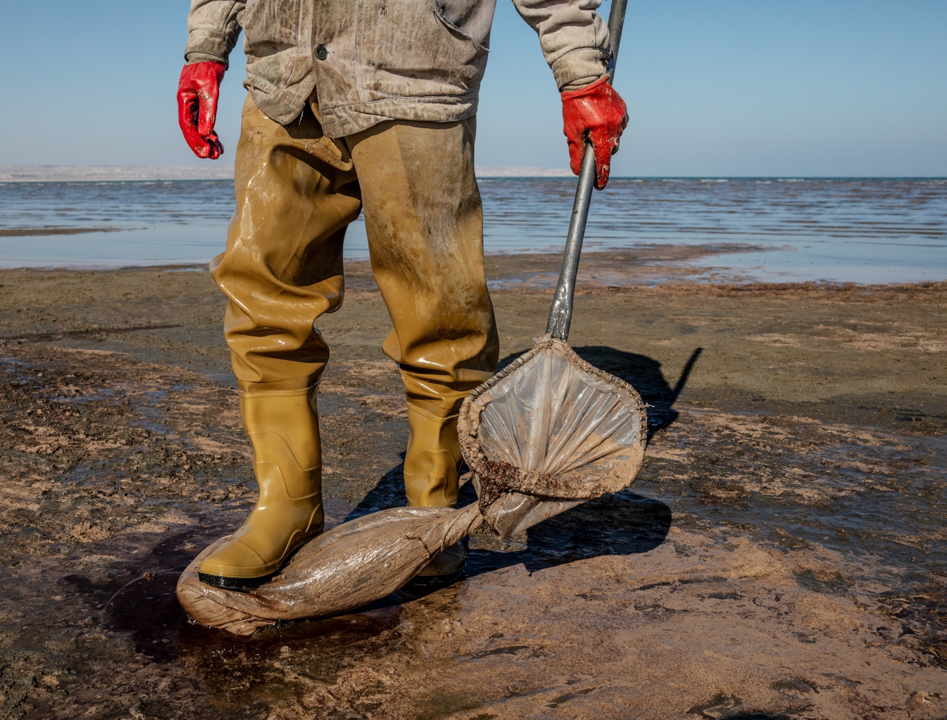 Low part of men's body in waders and hands in red glover holding a fish net. Right food on plastic bag.