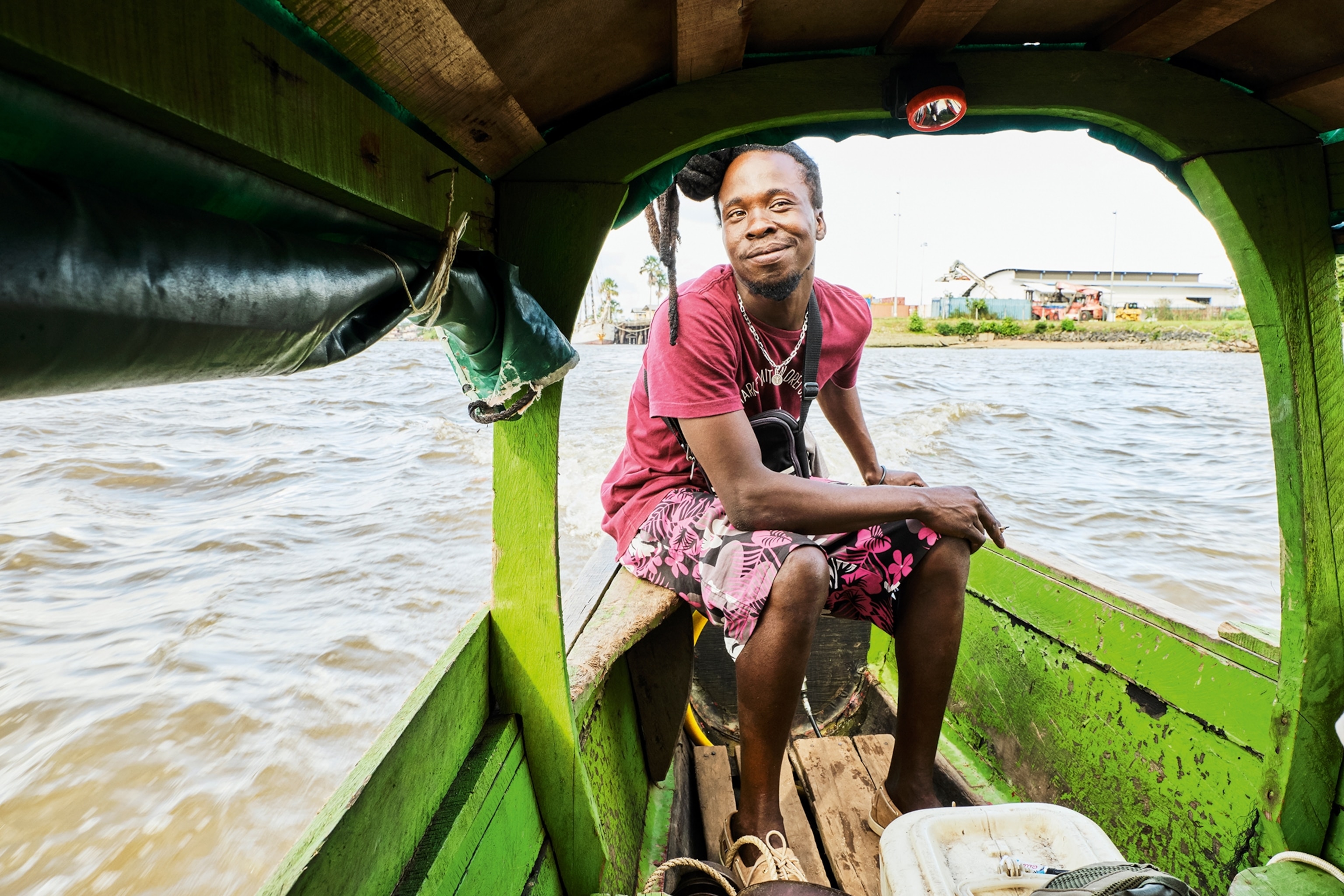 A smiling young local sitting at the rear of a small boat, steering it with one hand wearing a simple t-shirt and patterned swimming trunks.