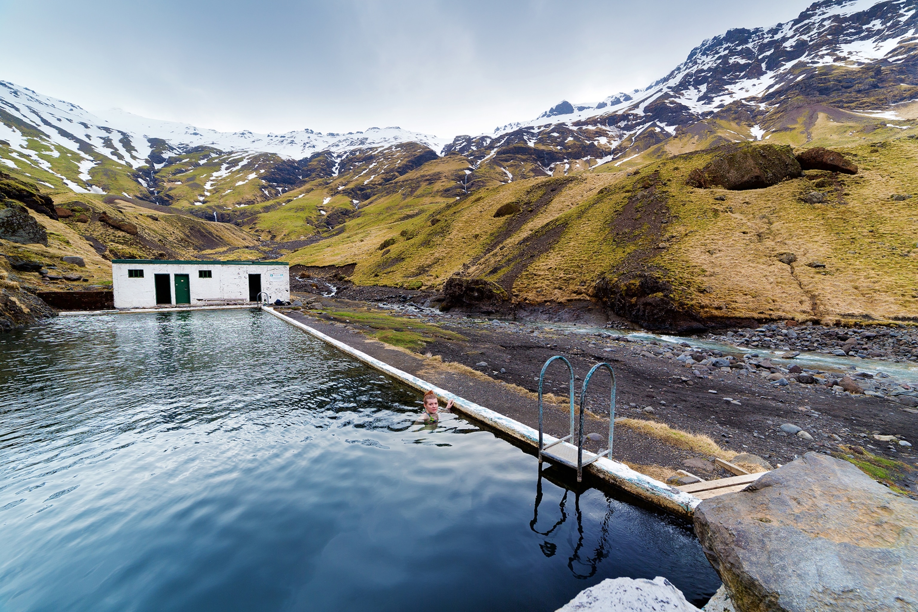 Pool filled with water surrounded by yellow-coloured hillsides and snowy mountain peaks with a white hut at the far end of the pool