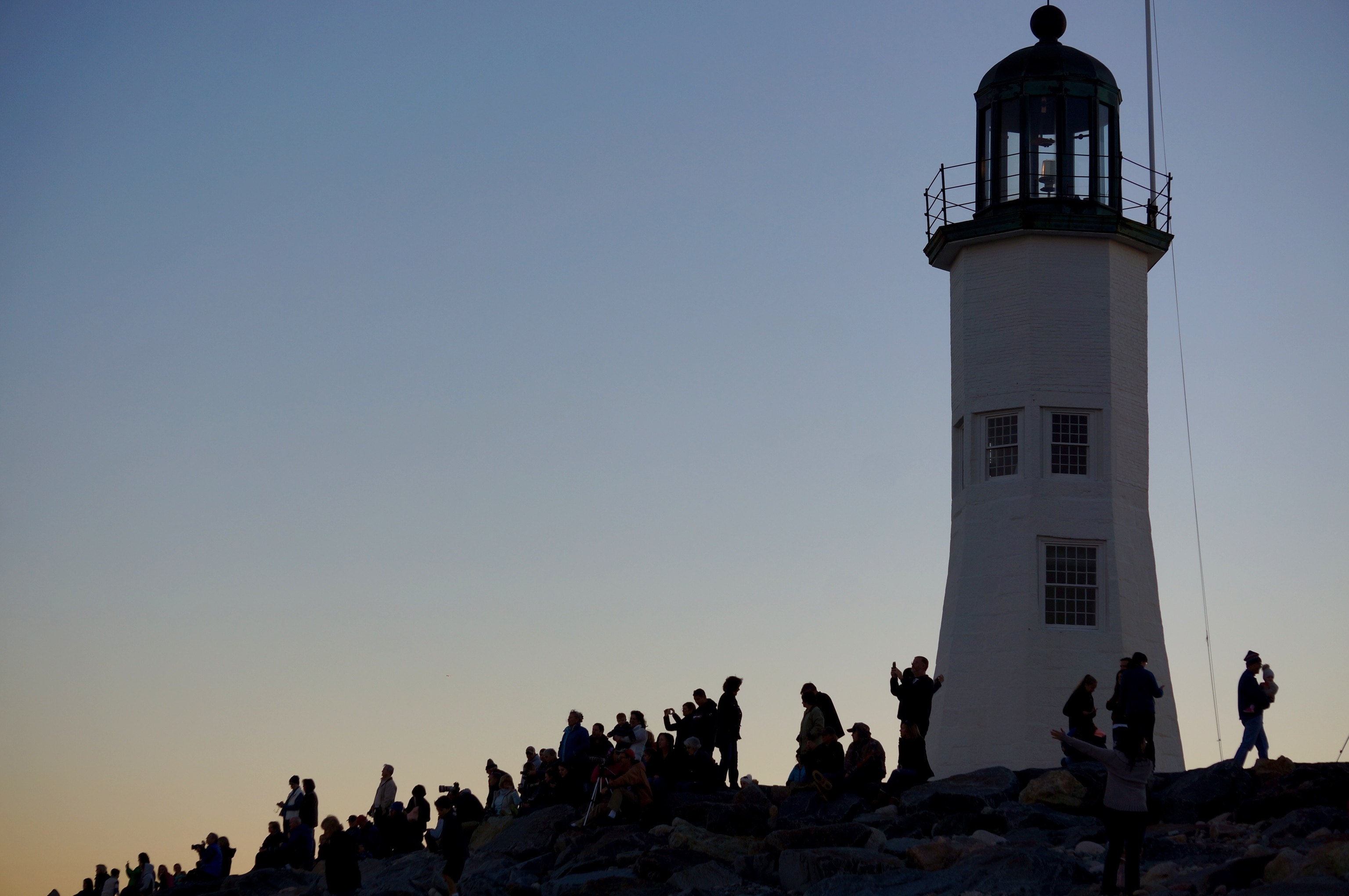 A large group of people, silhouetted by the sunset behind them, sand on rocks around the Scituate lighthouse