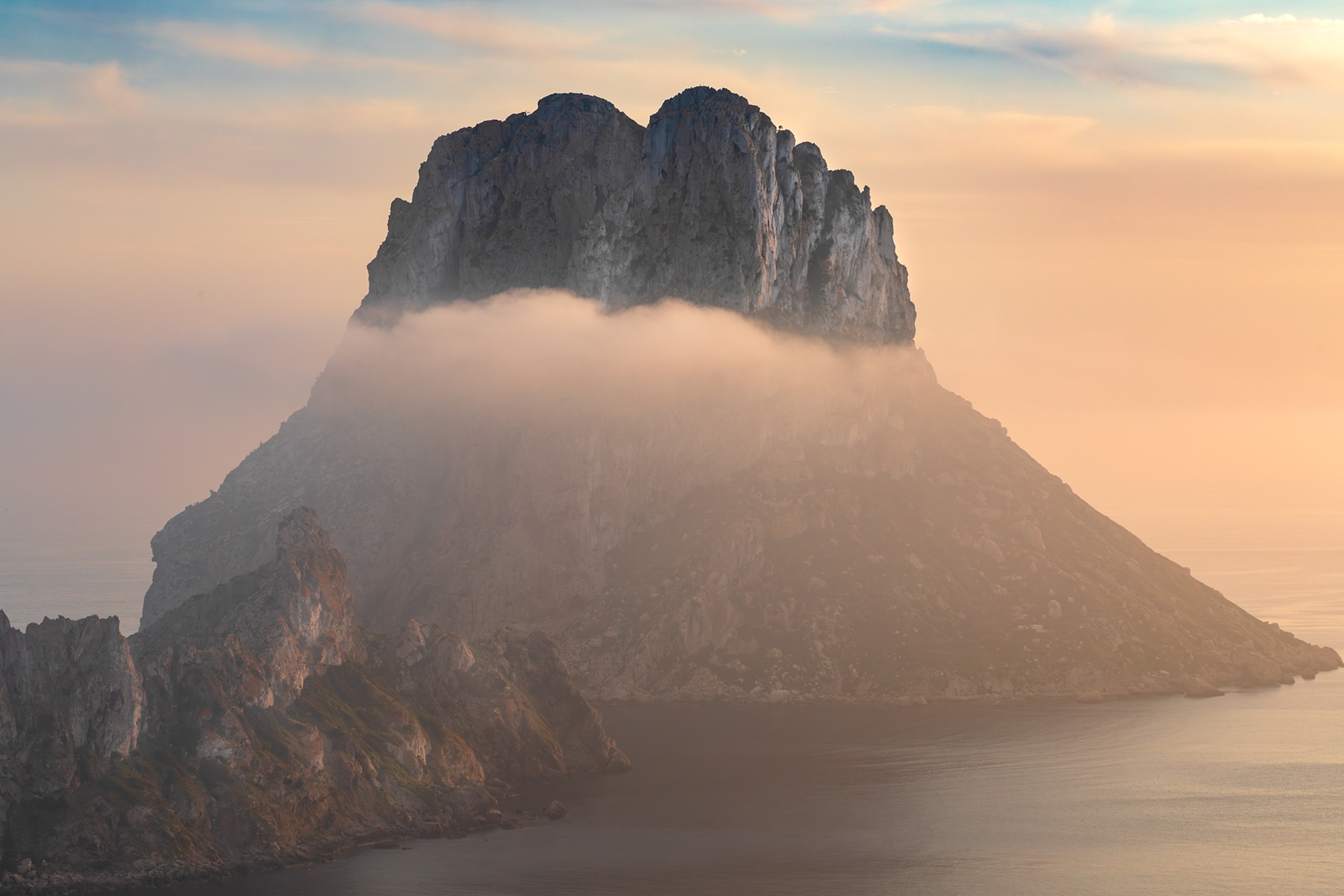 Misty mountain rises from the sea at sunset, with a cloud band encircling its peak.