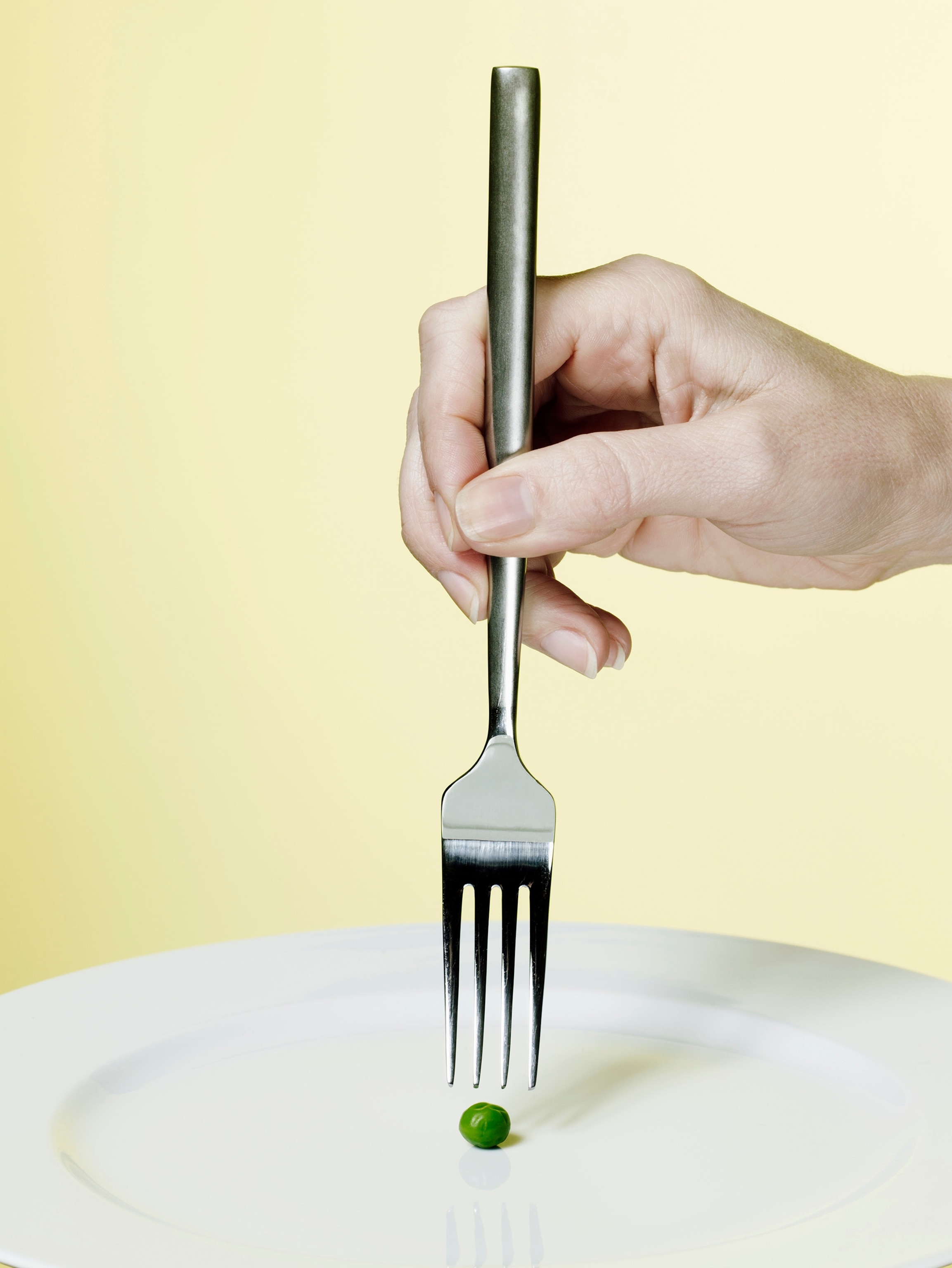 A close-up of a woman's hand holding a silver fork directly above a single green pea on a white plate against a pale yellow background.