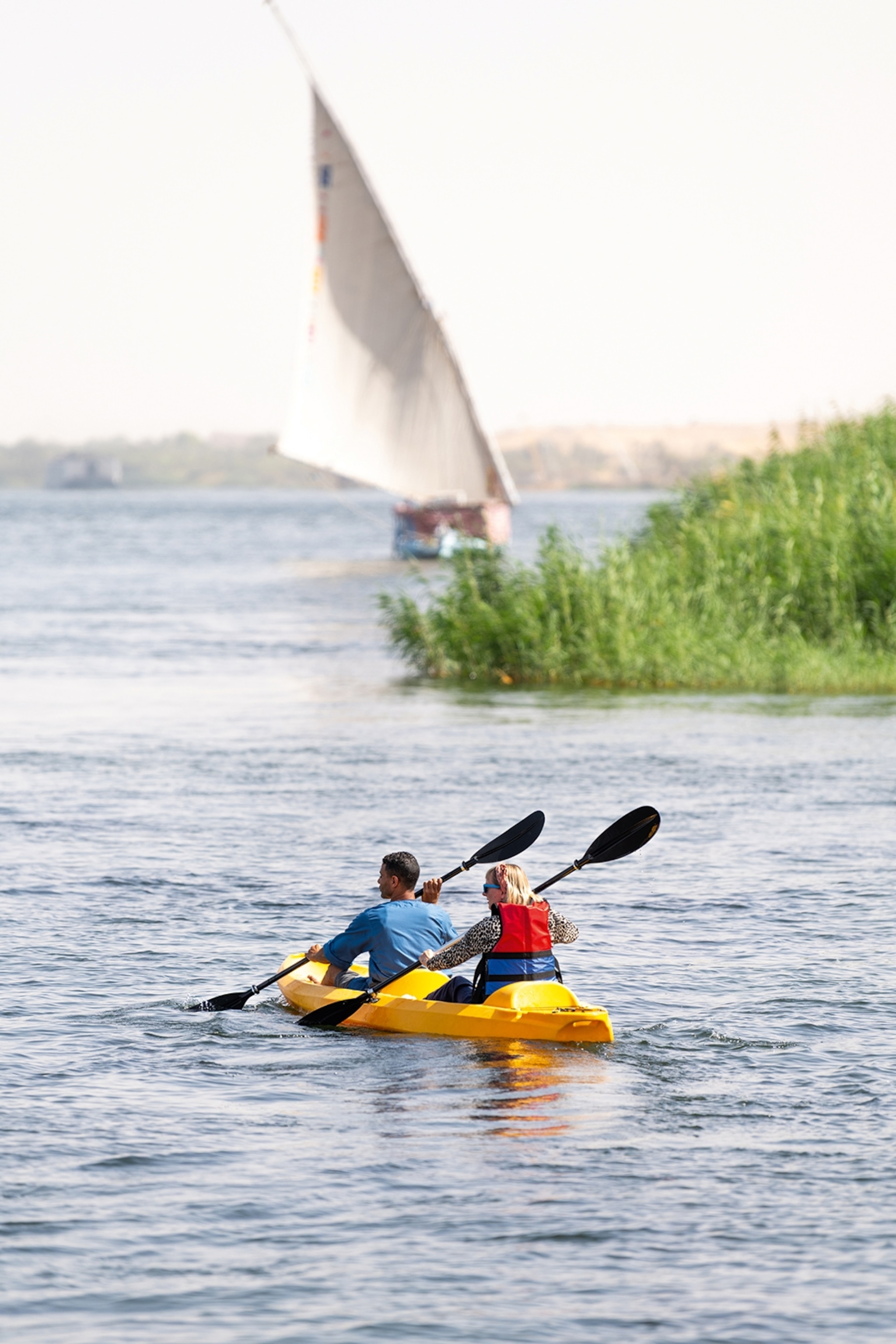 Two people in a kayak with a sailing boat in the distance.