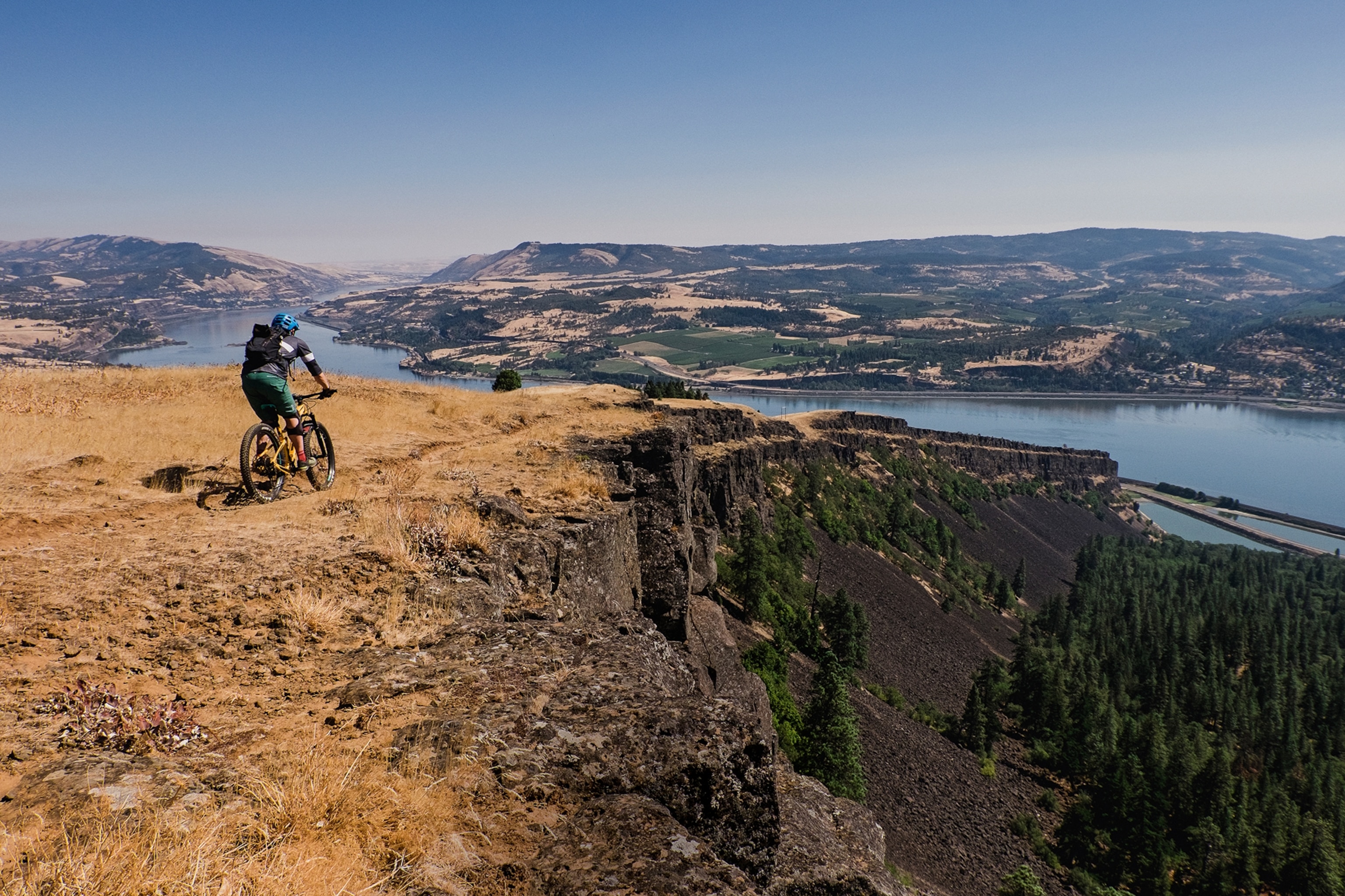 A biker on a mountain plateau with views over a wide river snaking through the hill-dotted landscape ahead.