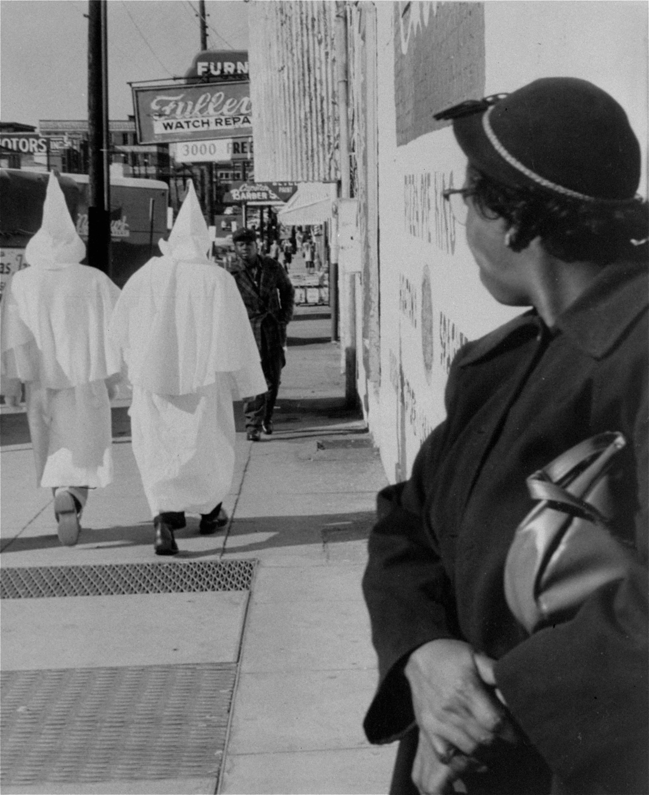 Klansmen in white robes are seen from behind as they walk down a street as an elderly African American woman watches on