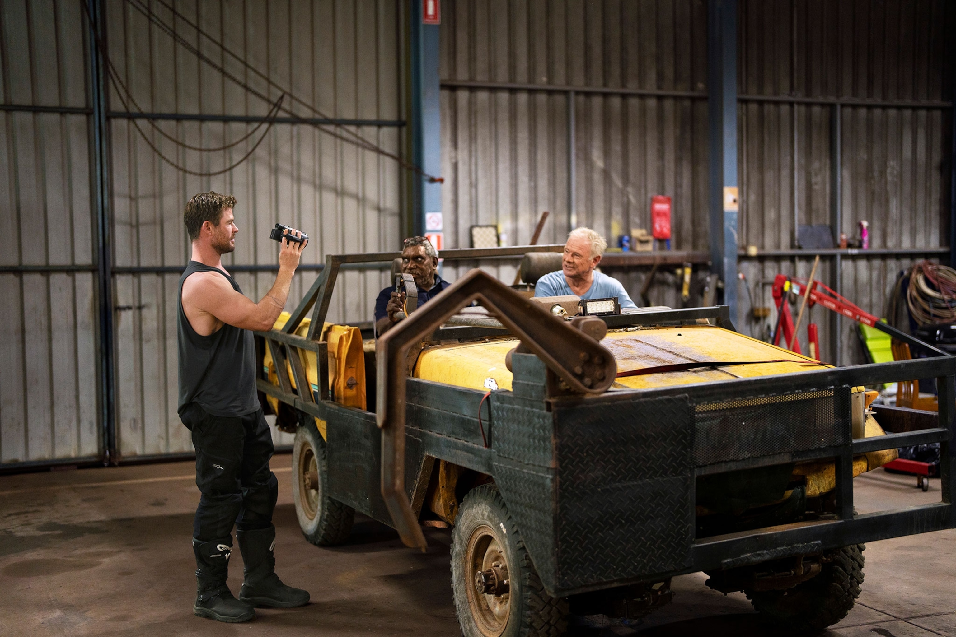 A man holds a camera at a yellow truck with two older men sitting inside one with one has a deep skin tone the other fair.