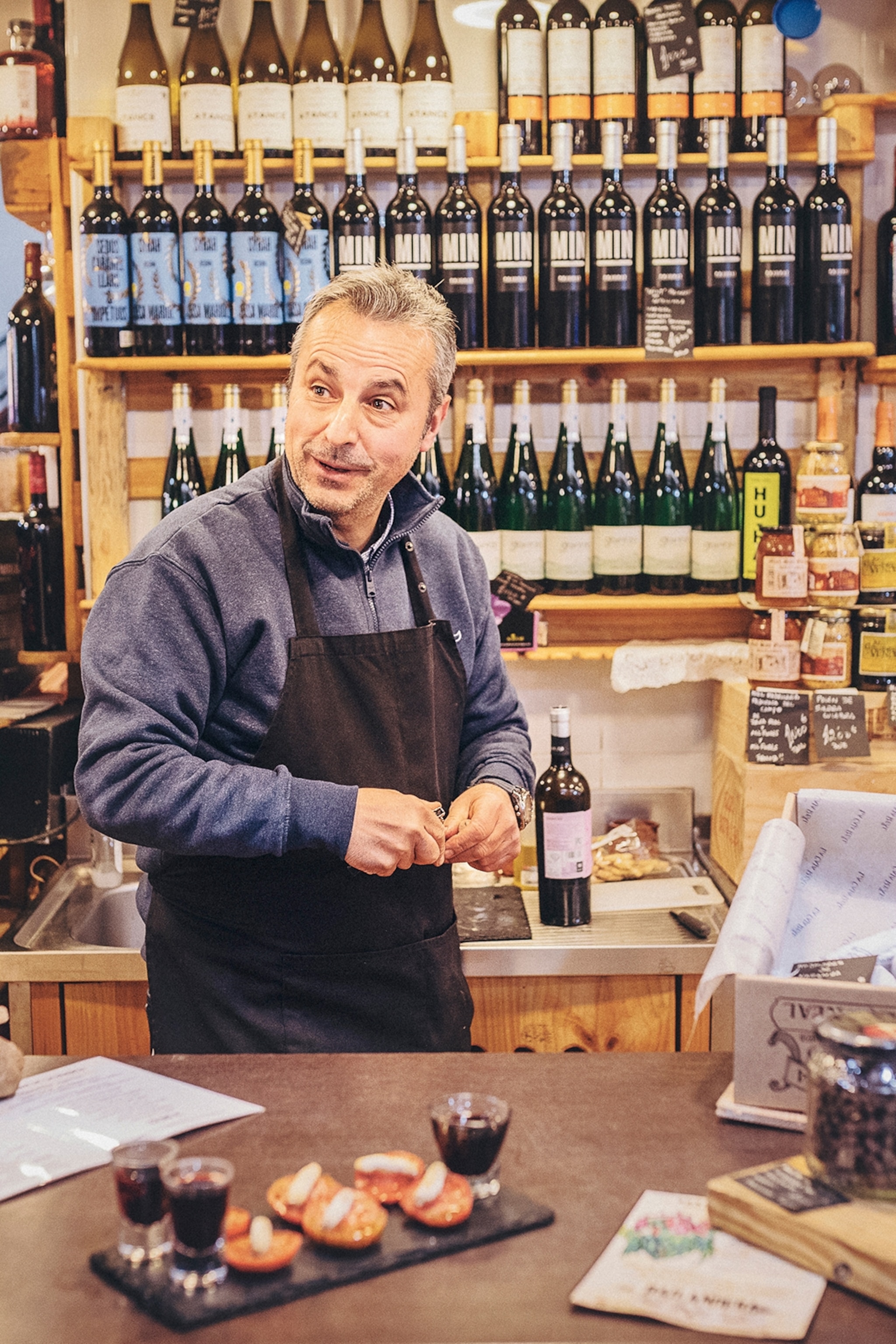 A male sale assistant in a delicatessen shop wearing an apron surrounded by bottles, jars and tins.