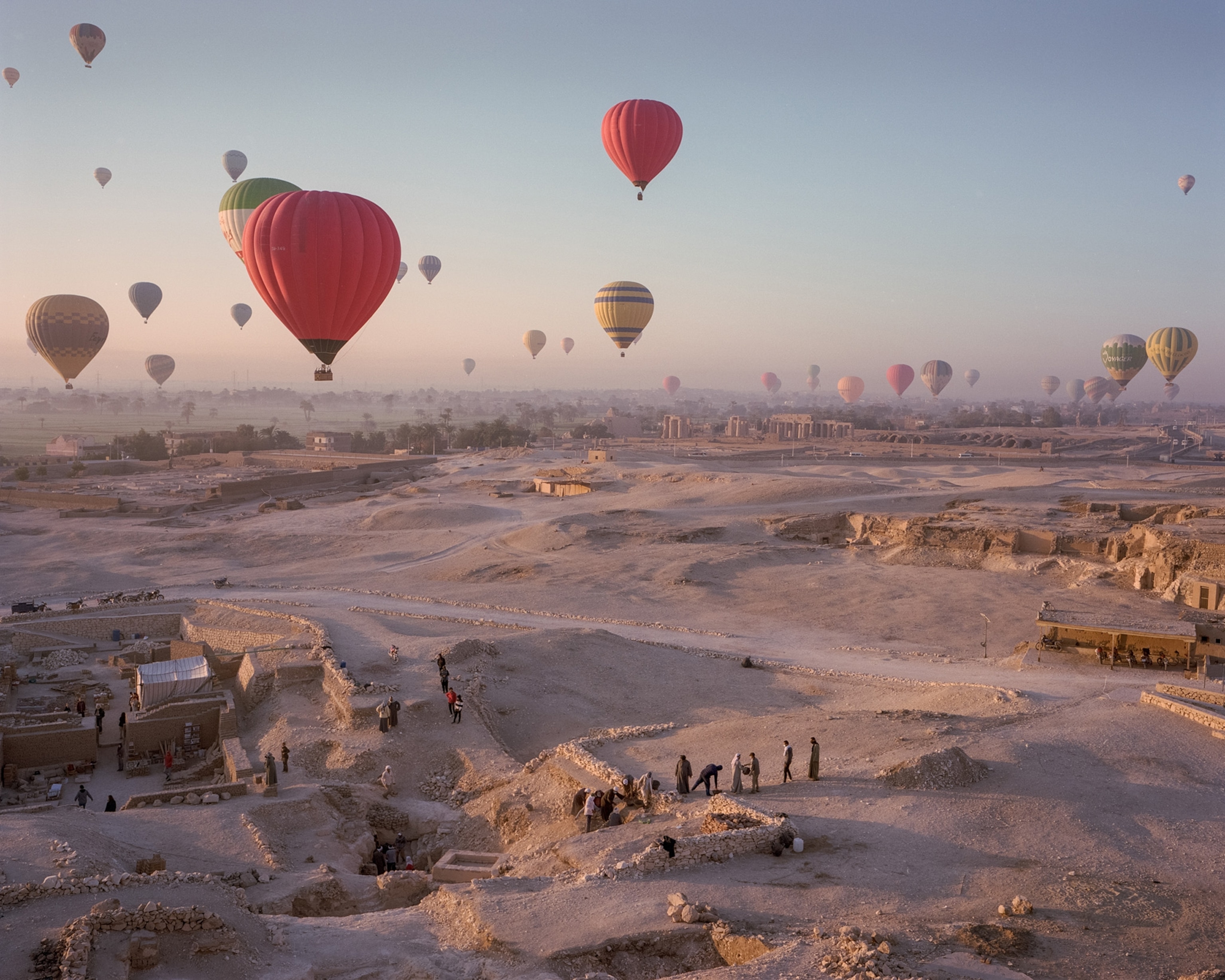 Dozens of hot air balloons fly over a vast, sandy excavation site.