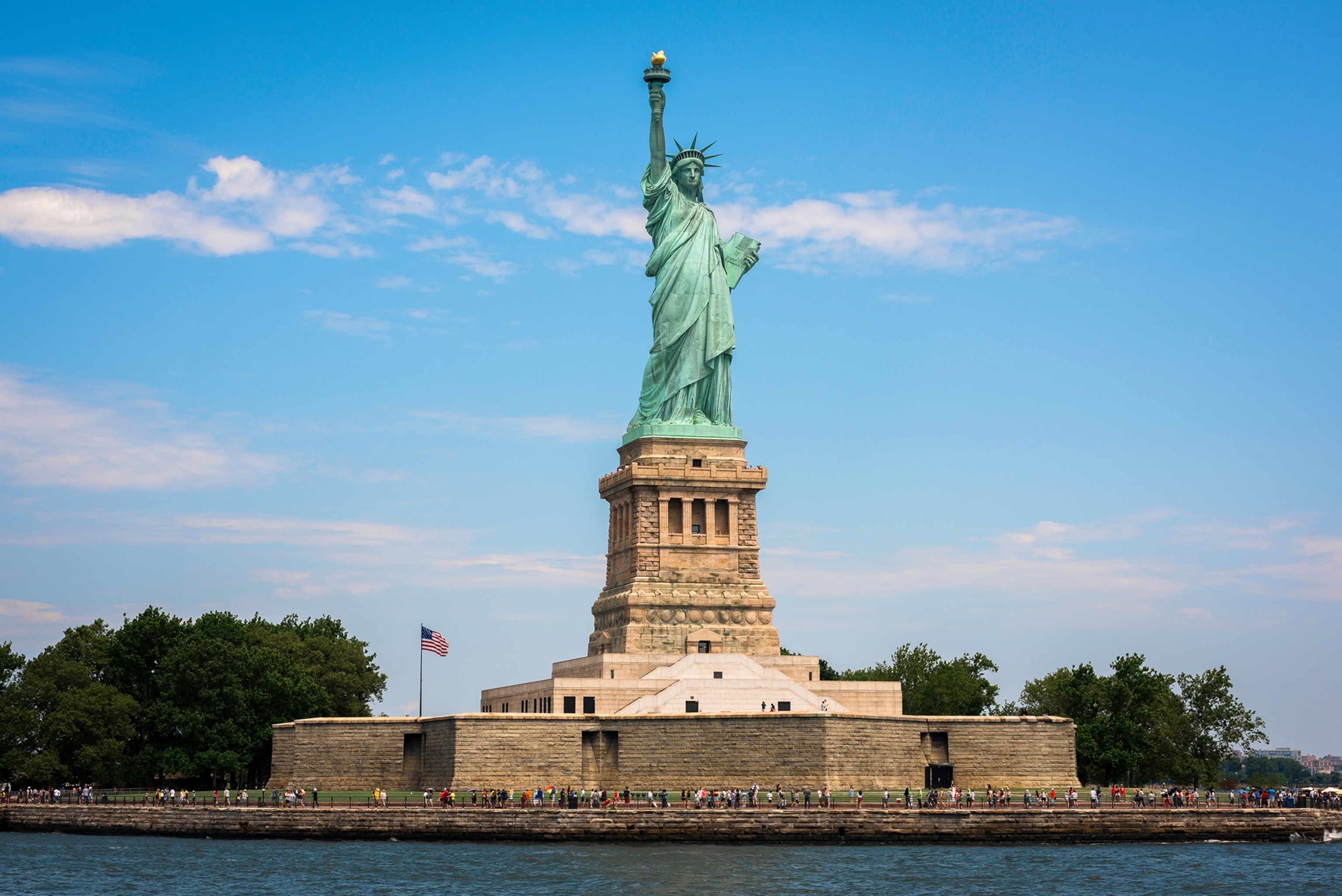A blue-green statue of a robed woman stands on a massive stone structure overlooking a bay