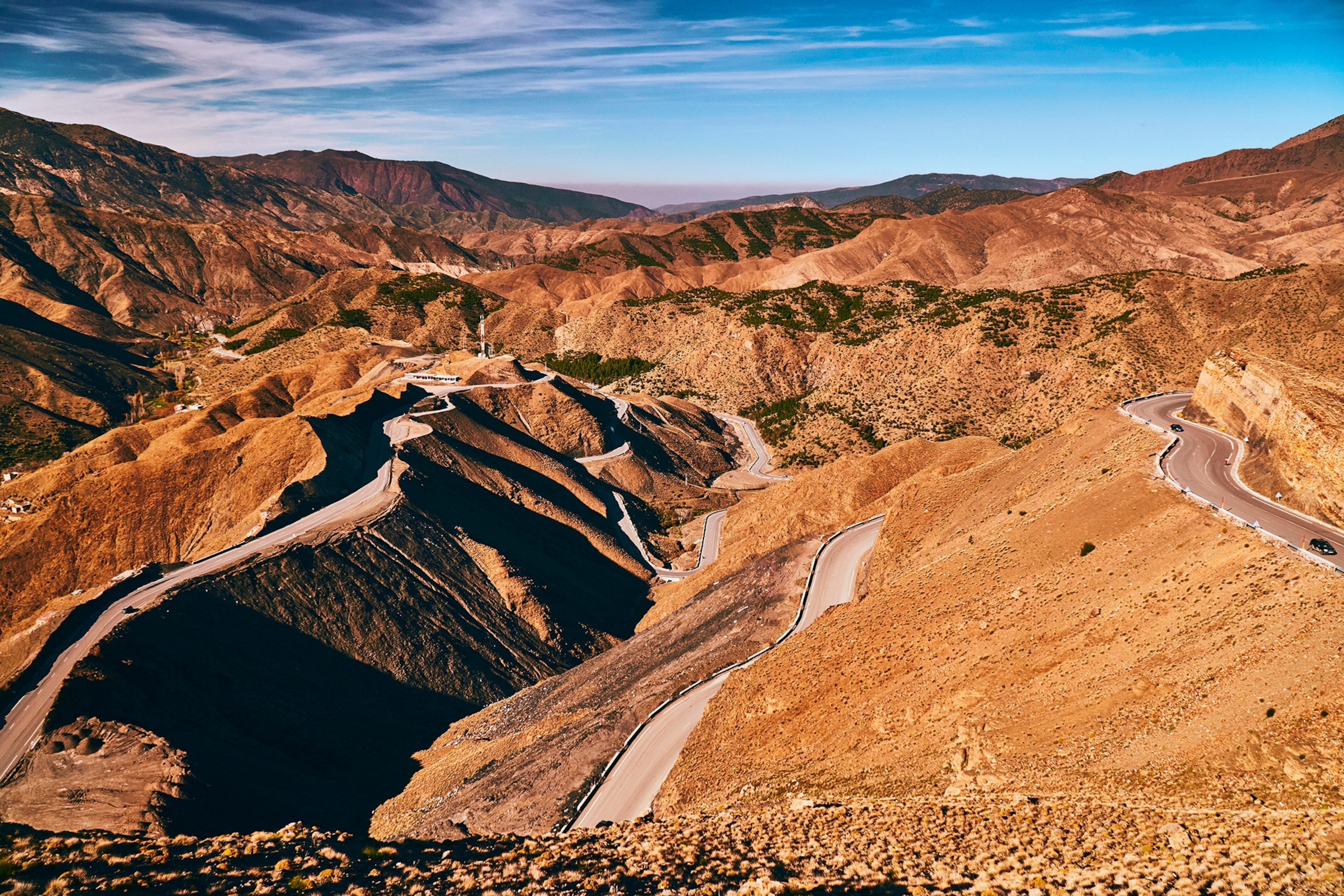 An aerial shot of a rugged mountain scape with a winding road leading in zig-zag across and around the hills.