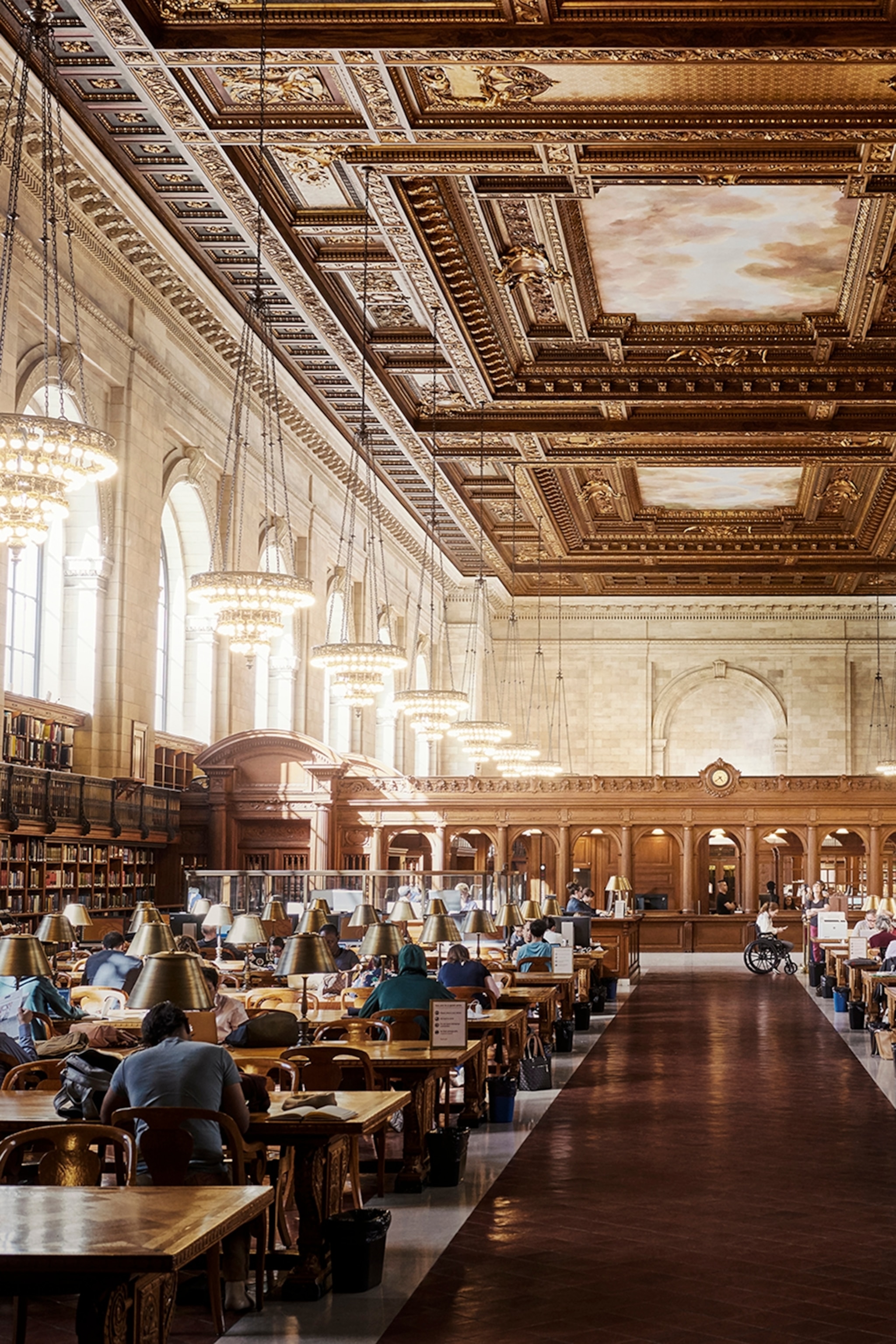 Interior of a grand building with people sat at wooden desks working while chandeliers hang above them