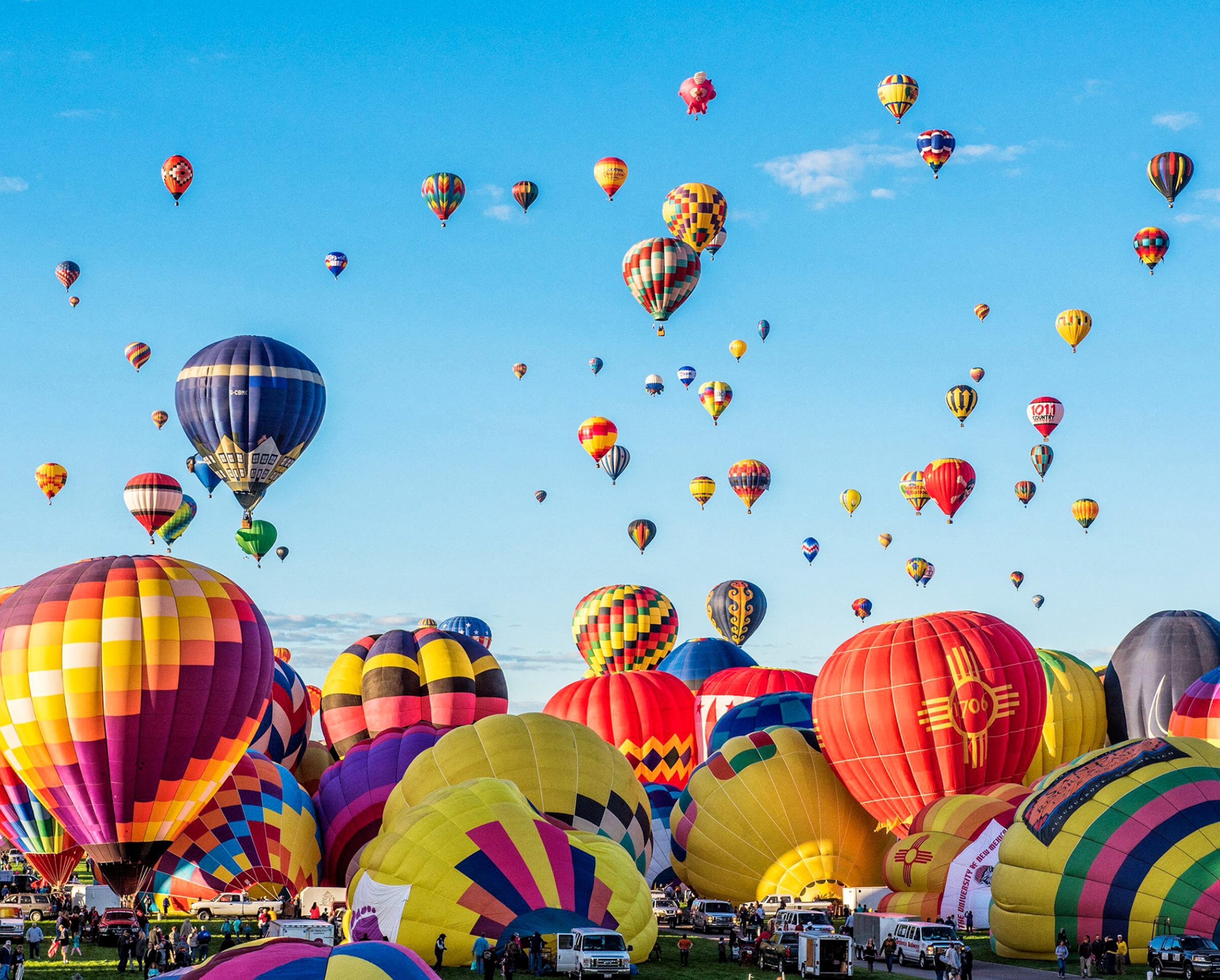 Dozens of large hot air balloons of various colors line the ground and are floating into the sky in the background. People are walking on the grass around and below the balloons.