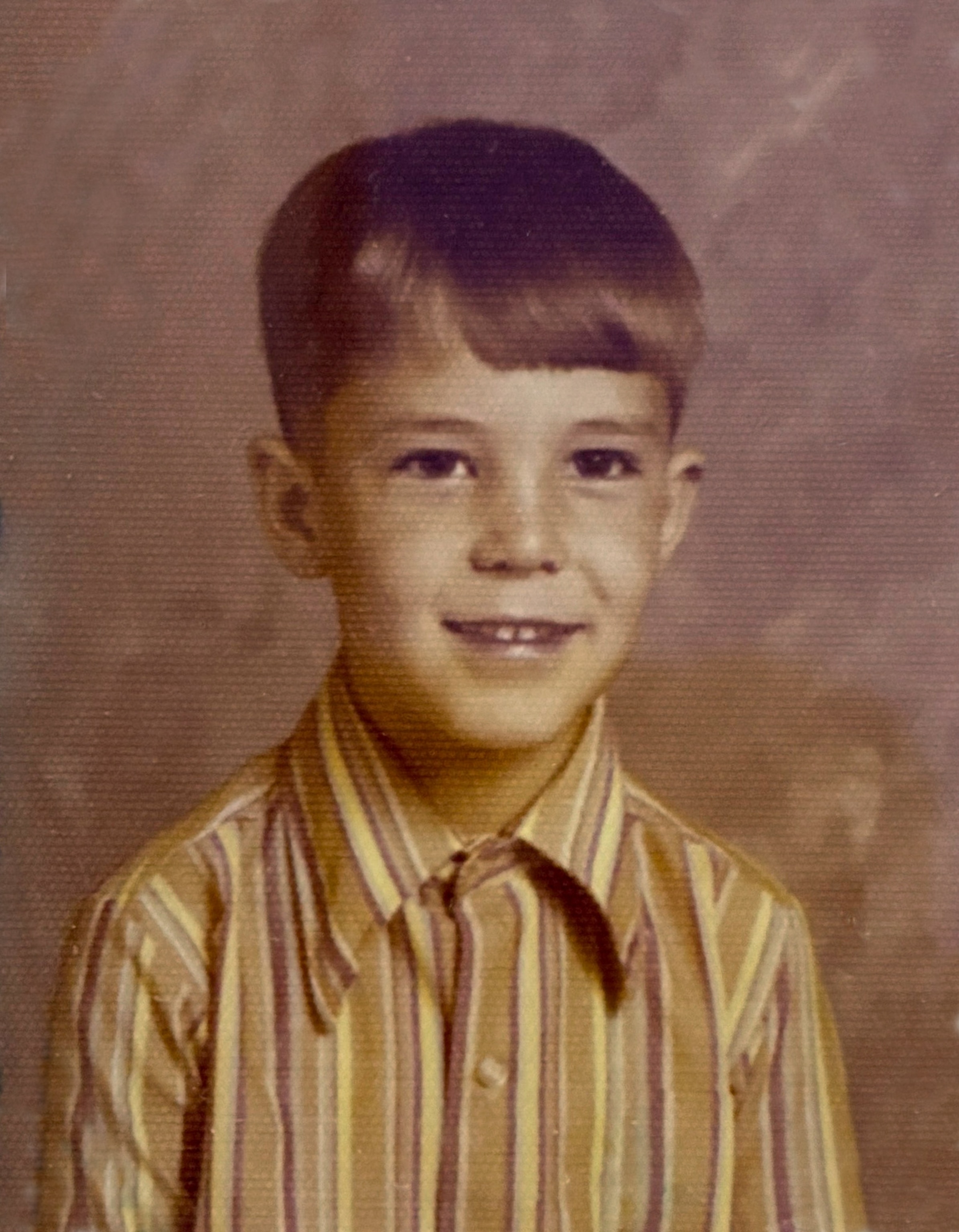A young boy smiles warmly in a vintage photo, wearing a striped brown and yellow shirt.