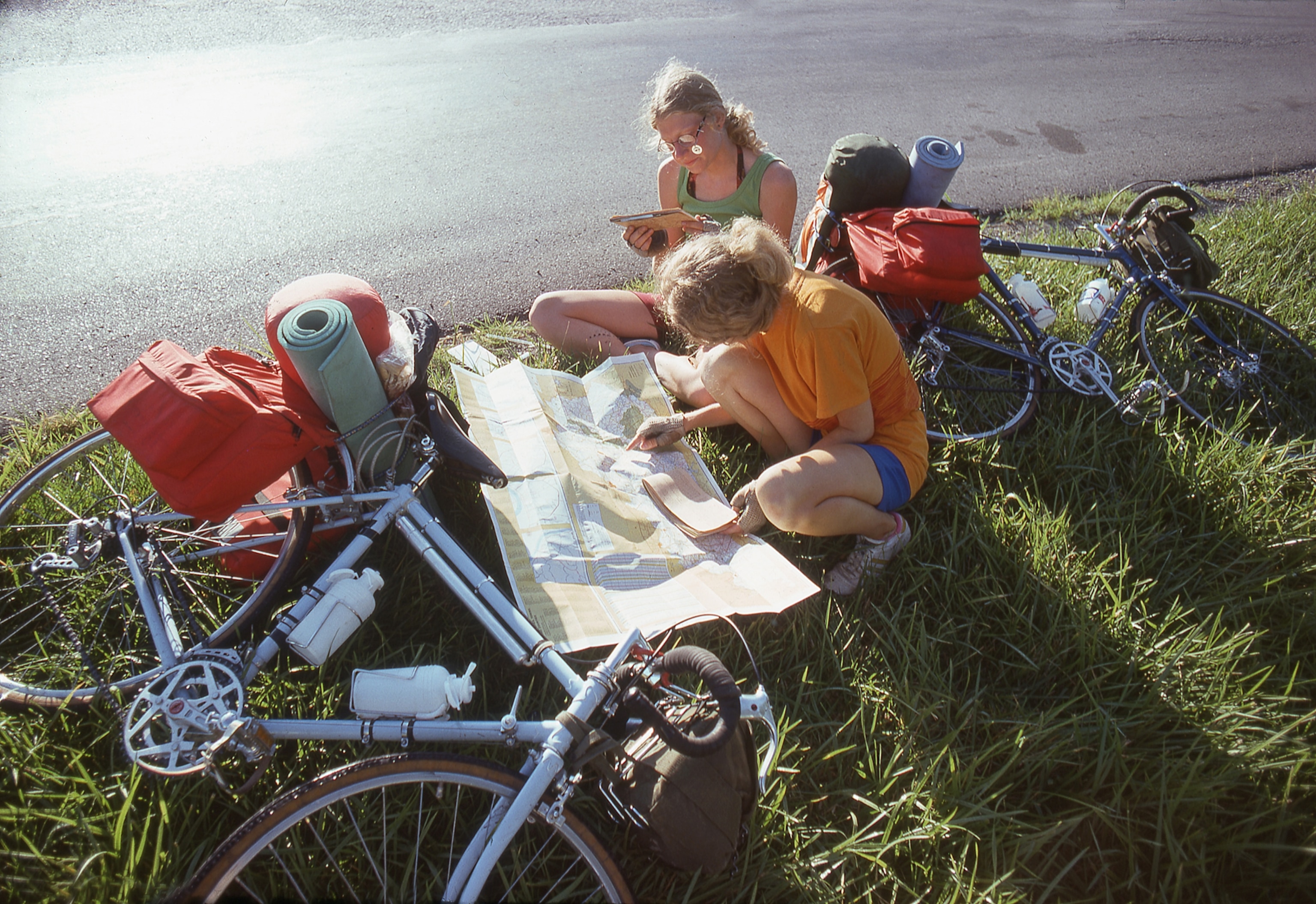 two young women sit on the side of the road reading a fold out paper mat next to their bicycles.