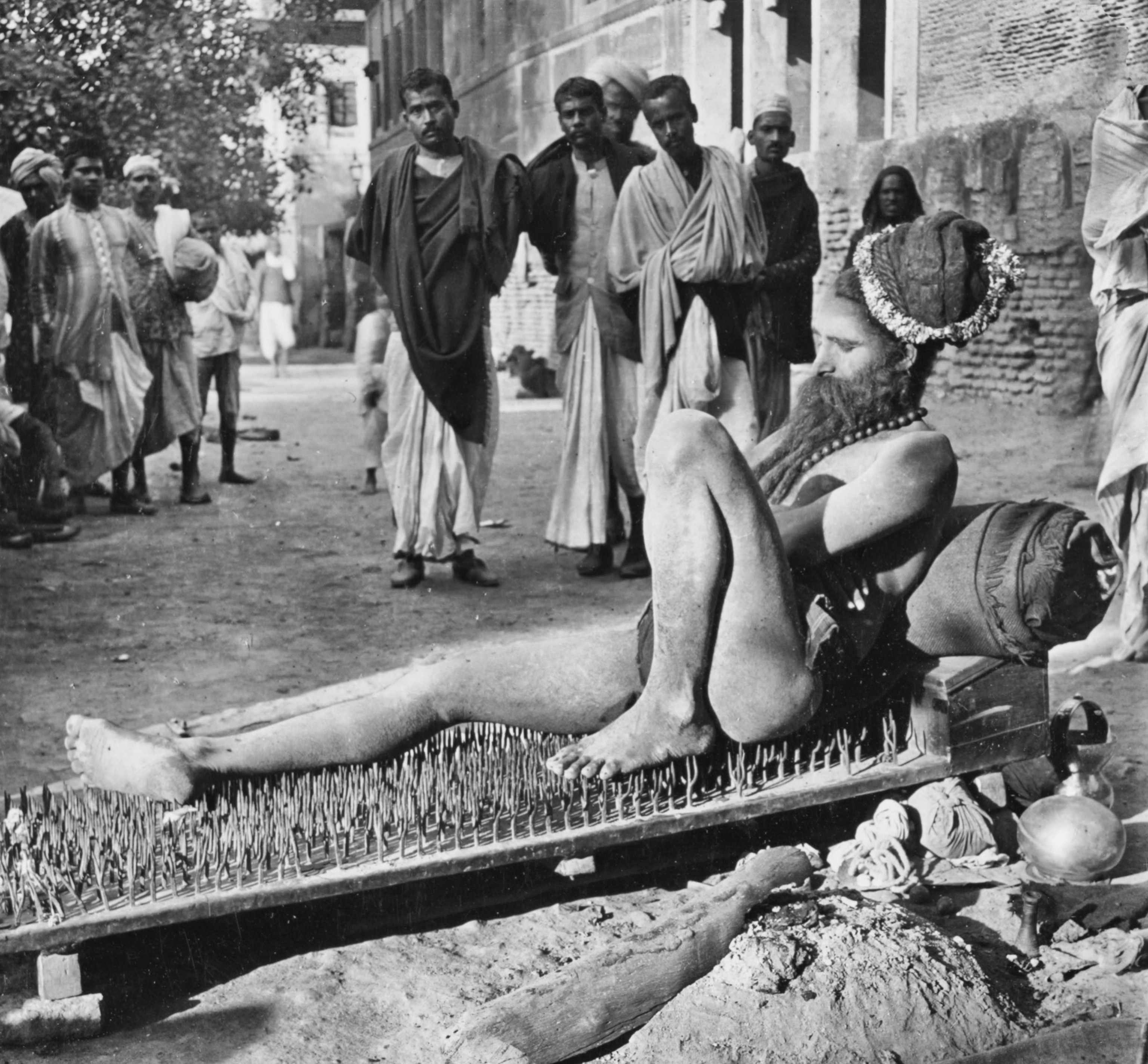 An Indian fakir resting atop a bed of nails as a gathering crowd looks on.