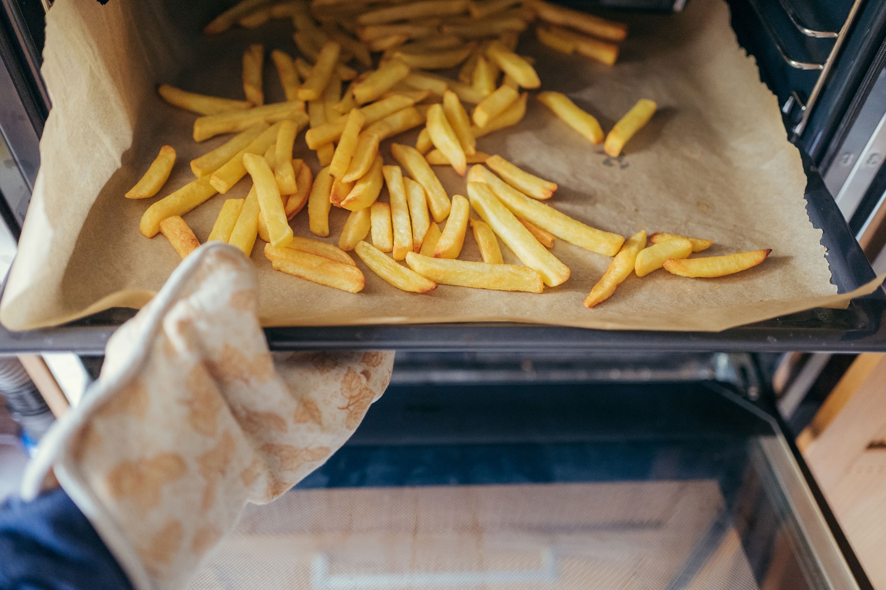 A hand using oven mitts takes a tray of french fries from the oven.