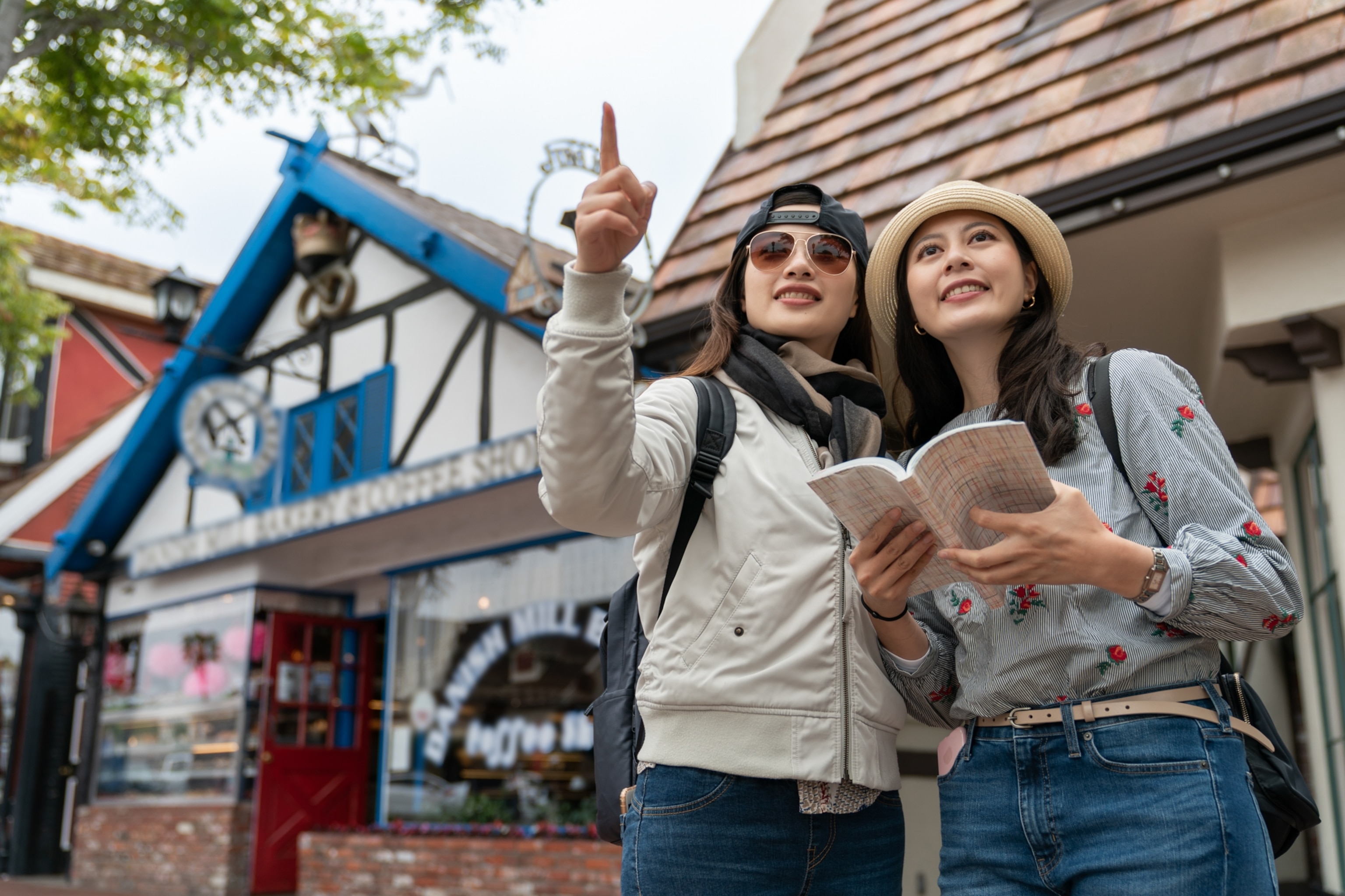 Two women stand outside a charming building. One points ahead, while the other holds a guidebook.
