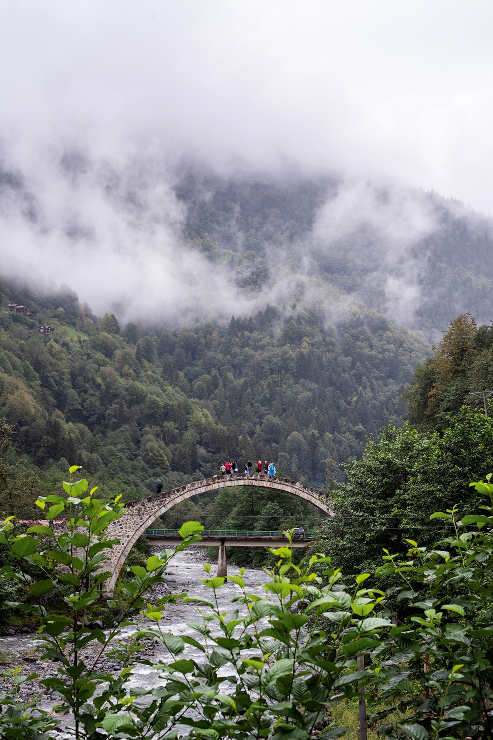 An arched stone bridge leading over a rocky river at the foot of a mountain hidden behind fog.