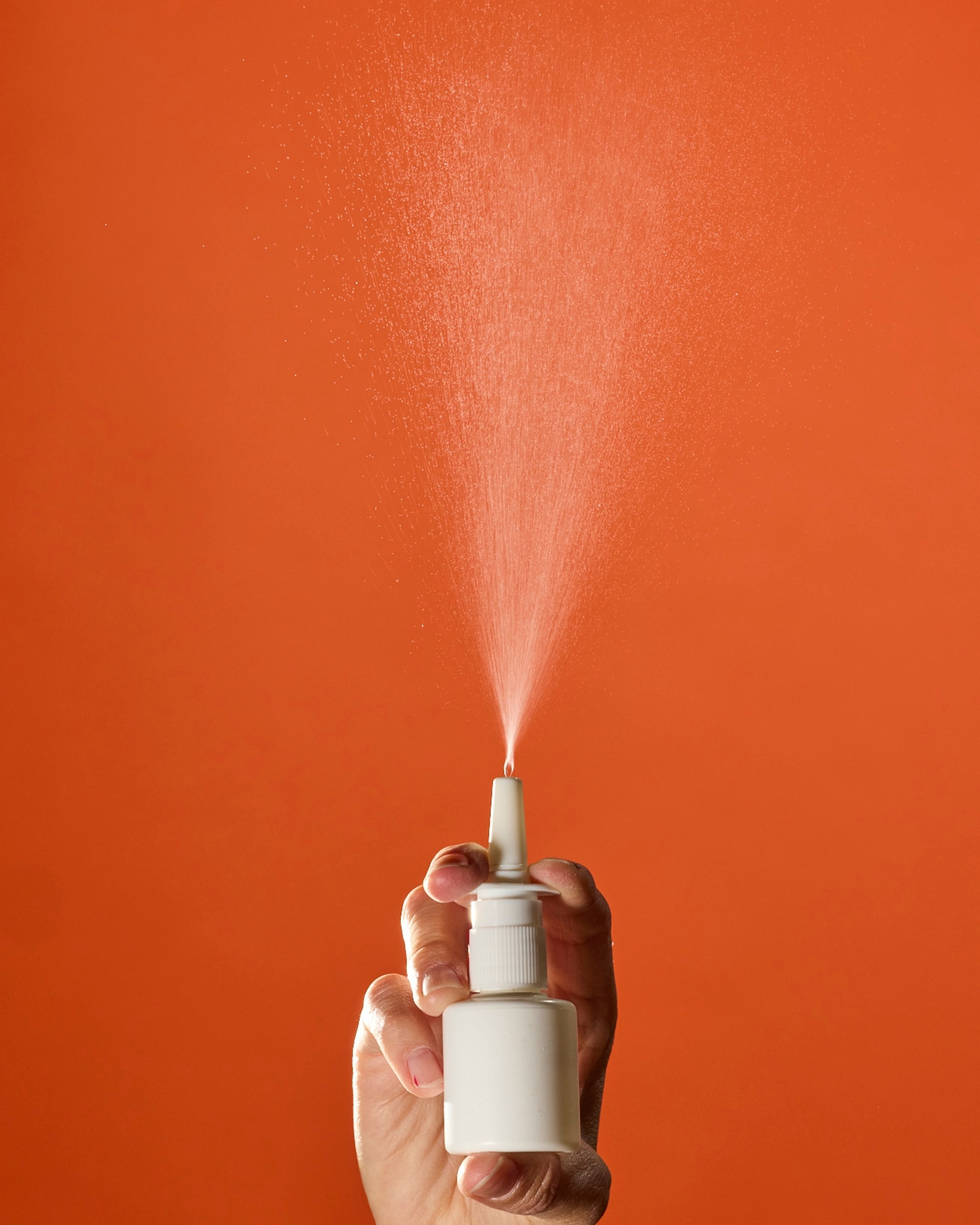 A person sprays an unlabeled white nasal spray against a burnt orange background