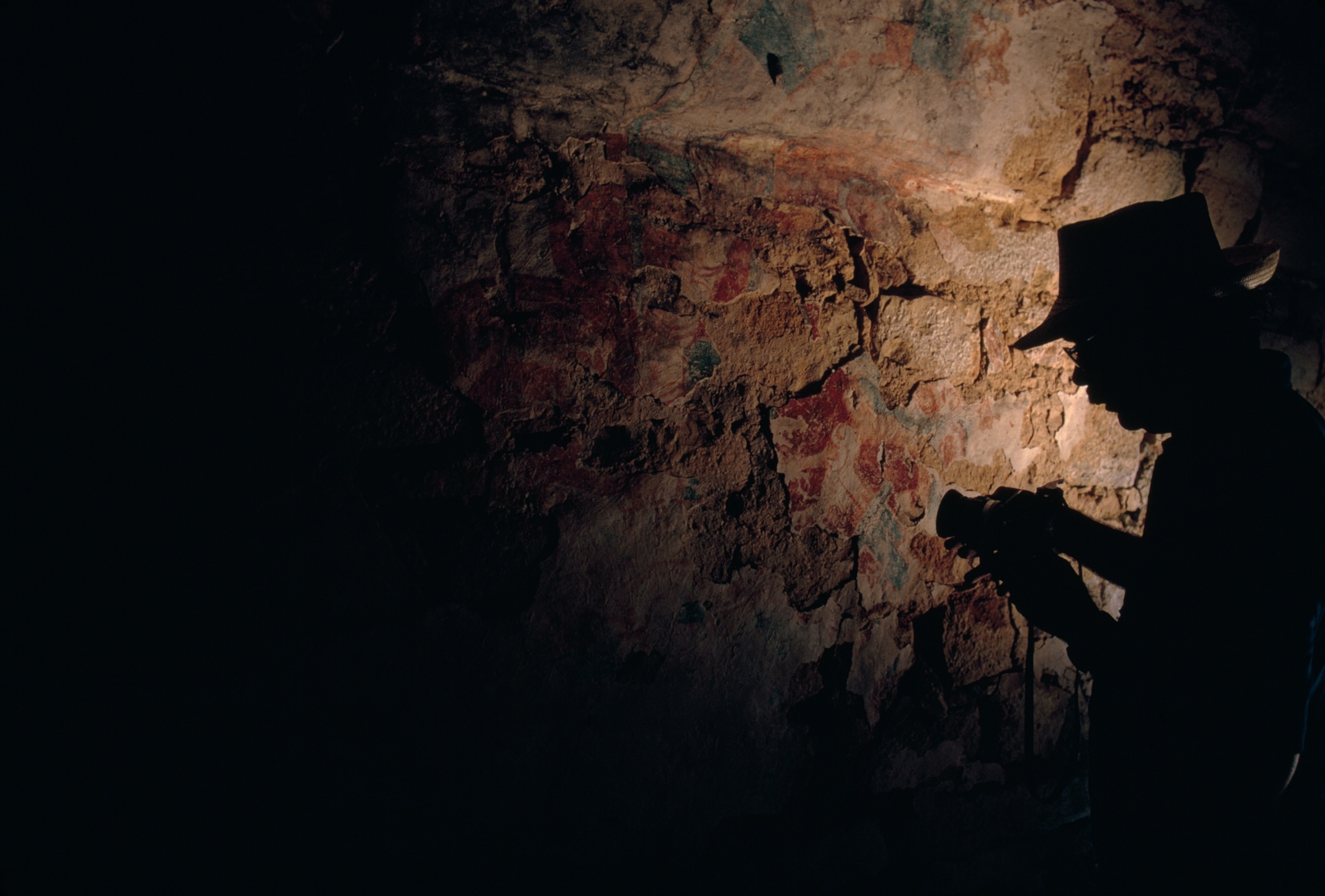 Silhouette of a person with a hat photographing a dimly lit, textured stone wall covered with ancient cave art.