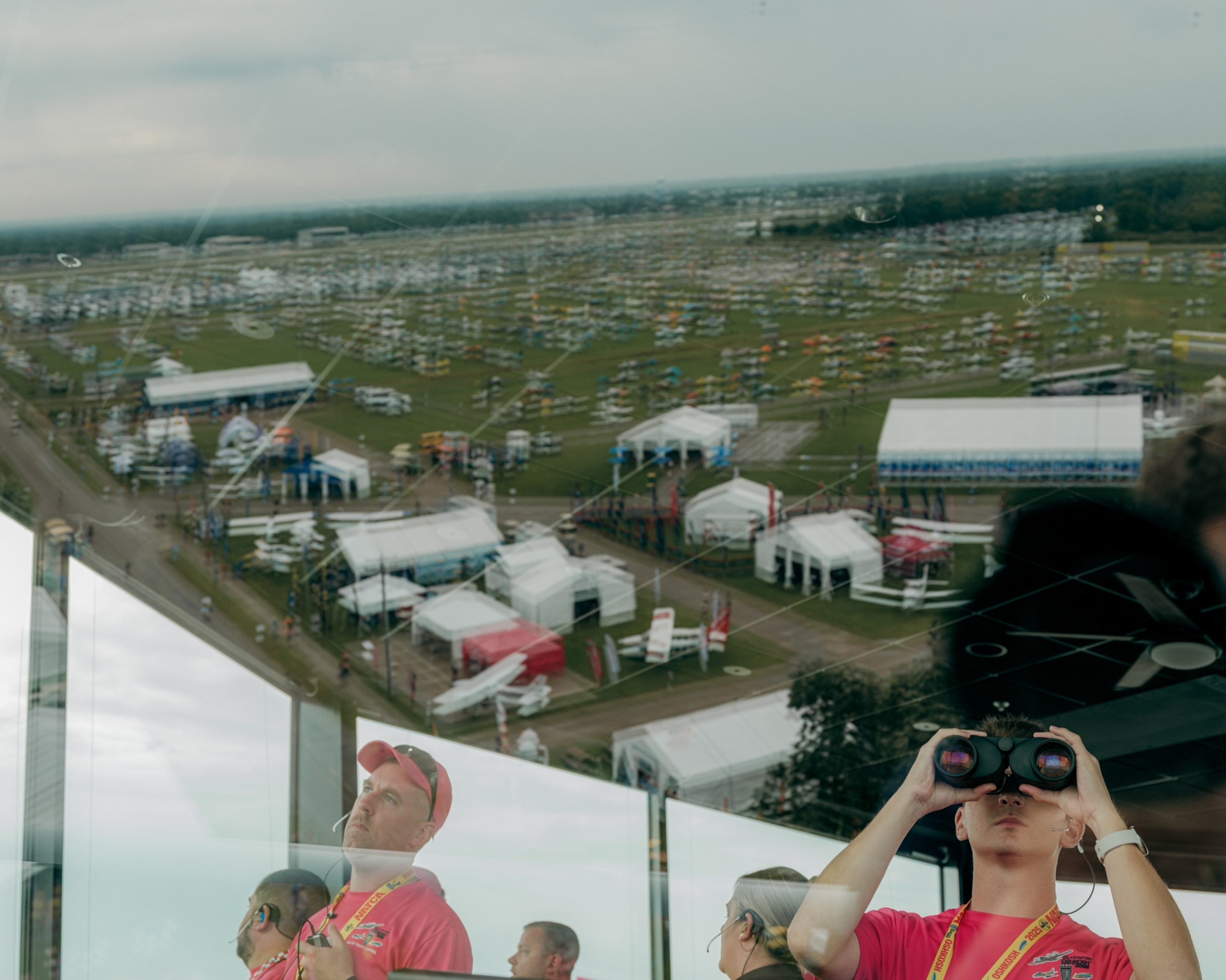 Aerial view reflected in glass of a crowded outdoor event with tents and vehicles. Inside the glass, a person looks through binoculars, wearing a bright pink shirt