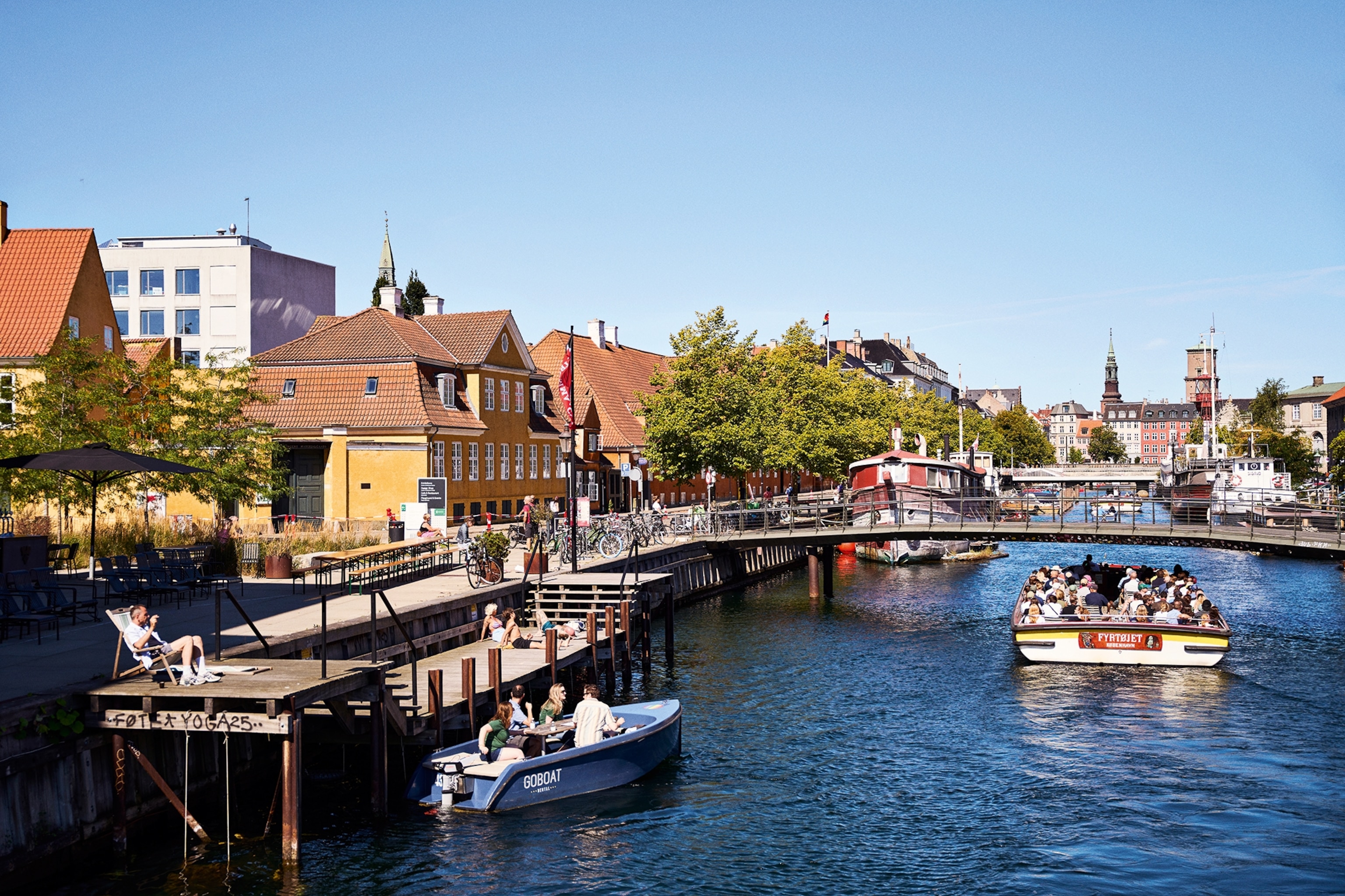 A harbour front with small boats taking off from either show, city buildings rising into view in the background.