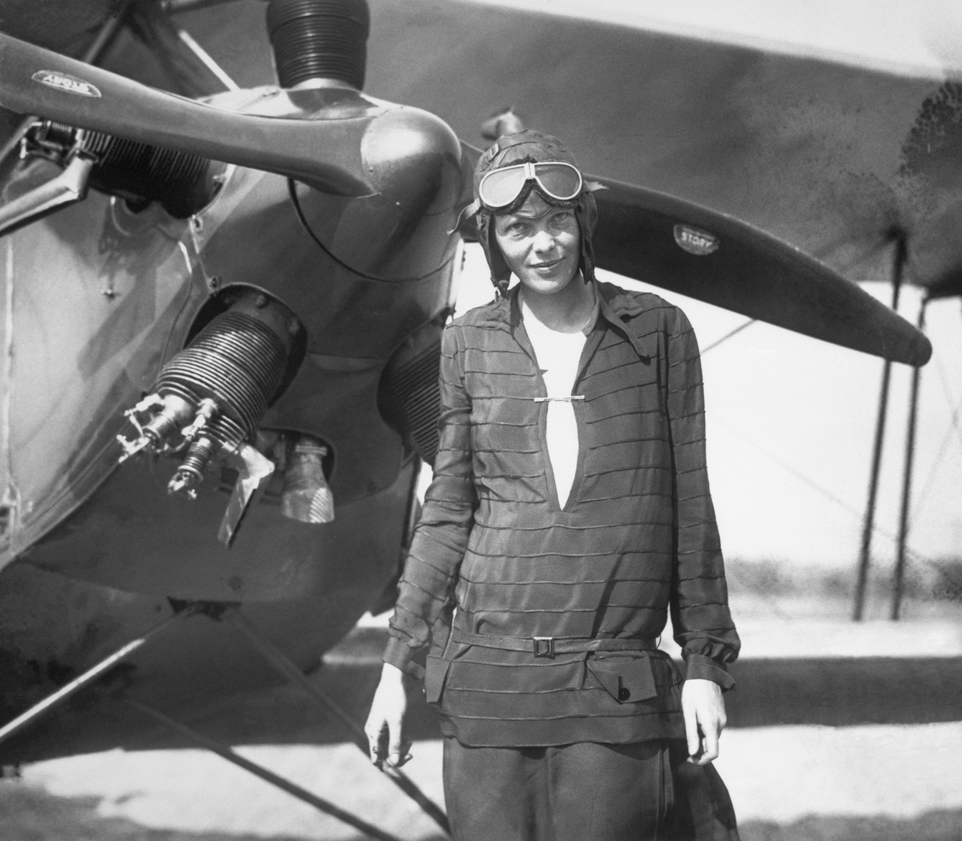 Black and white historical photo of a woman in a aviator's cap standing in front of a propeller plane