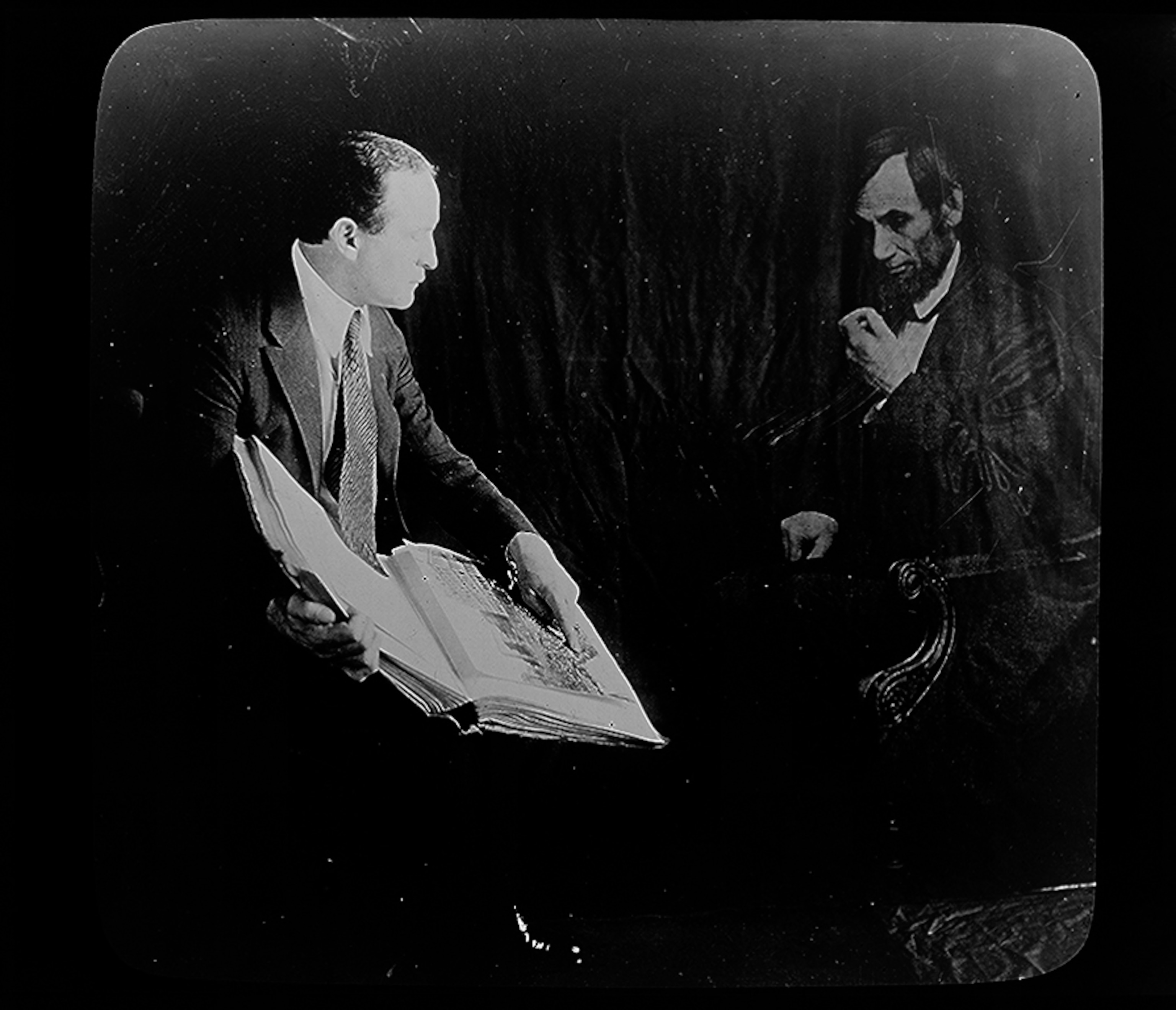 A man holds a book and as a figure with sideburns looks from the shadow.