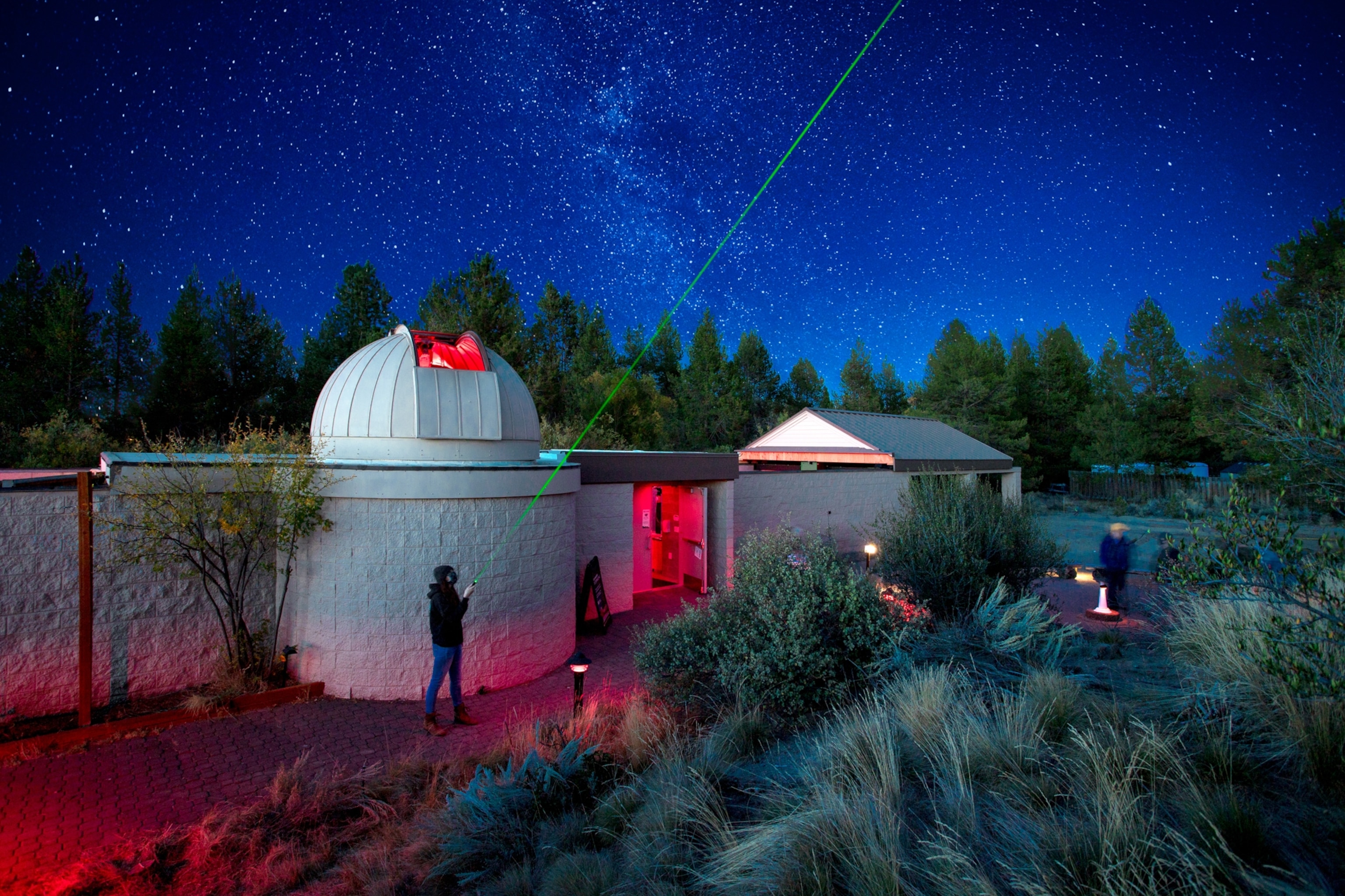 Observatory under a starry night sky with a green laser pointing upwards. A person stands nearby