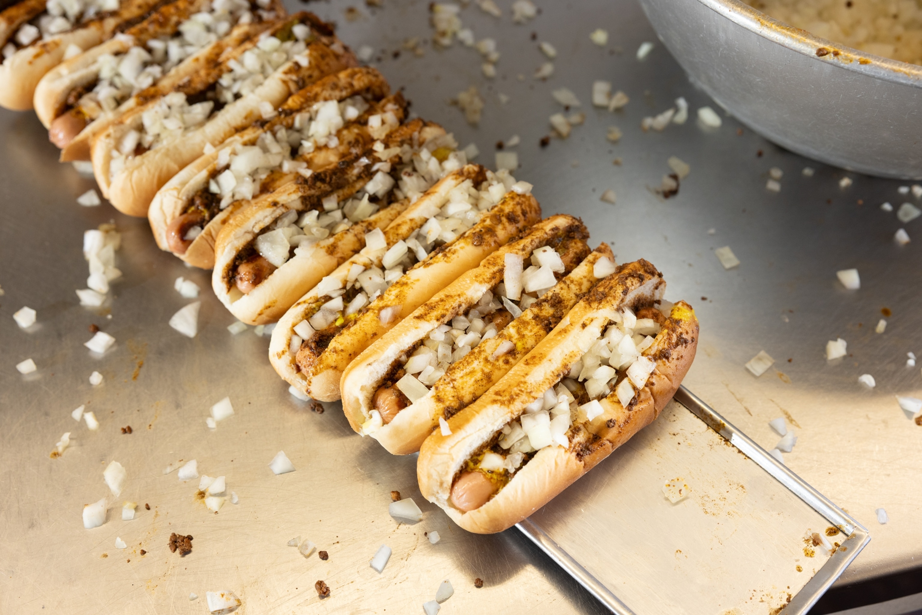 A row of hot dogs in buns topped with onions and chili on a metal table top