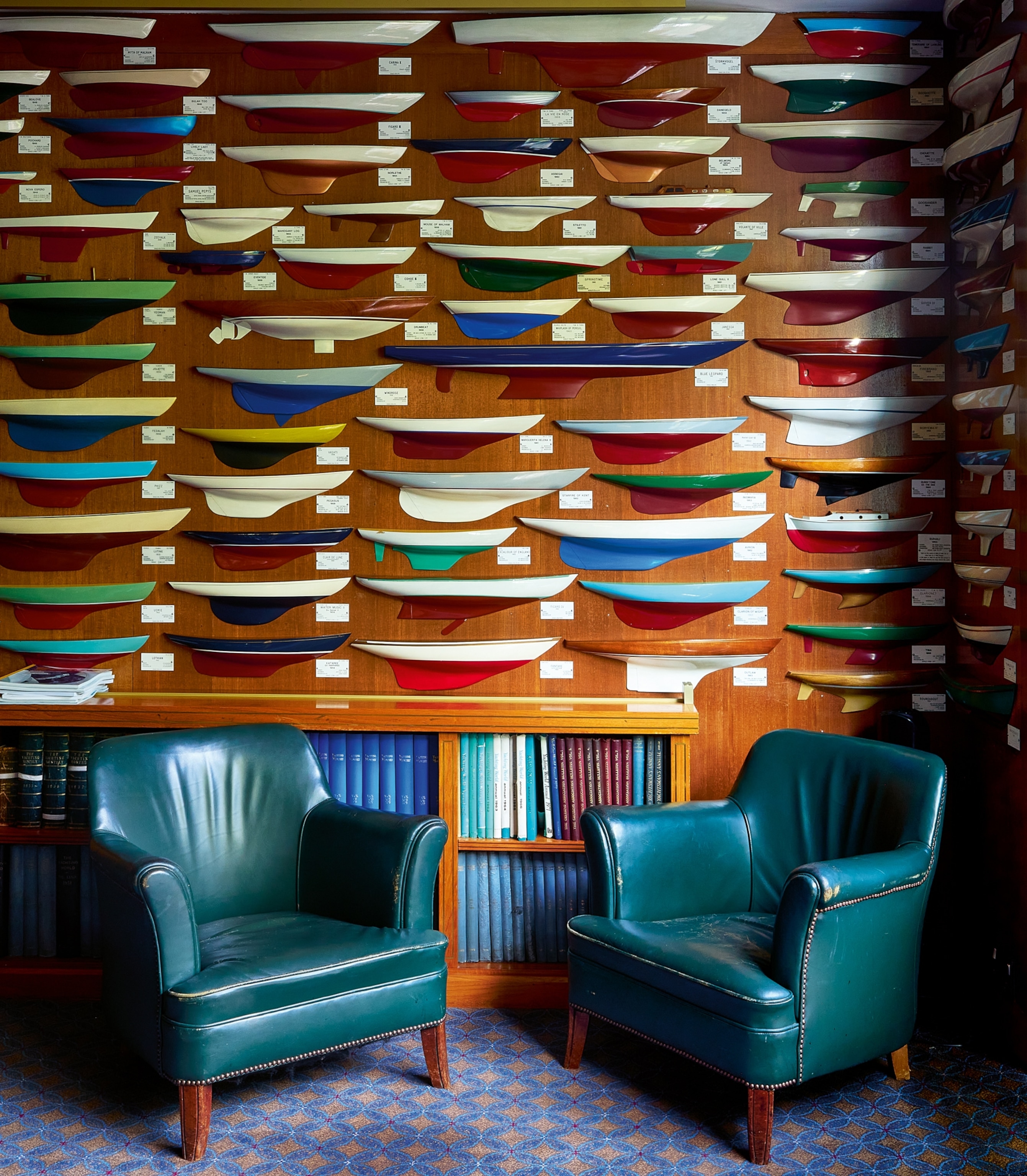 Room with two green armchairs, a wooden wall displaying colorful model ship hulls.