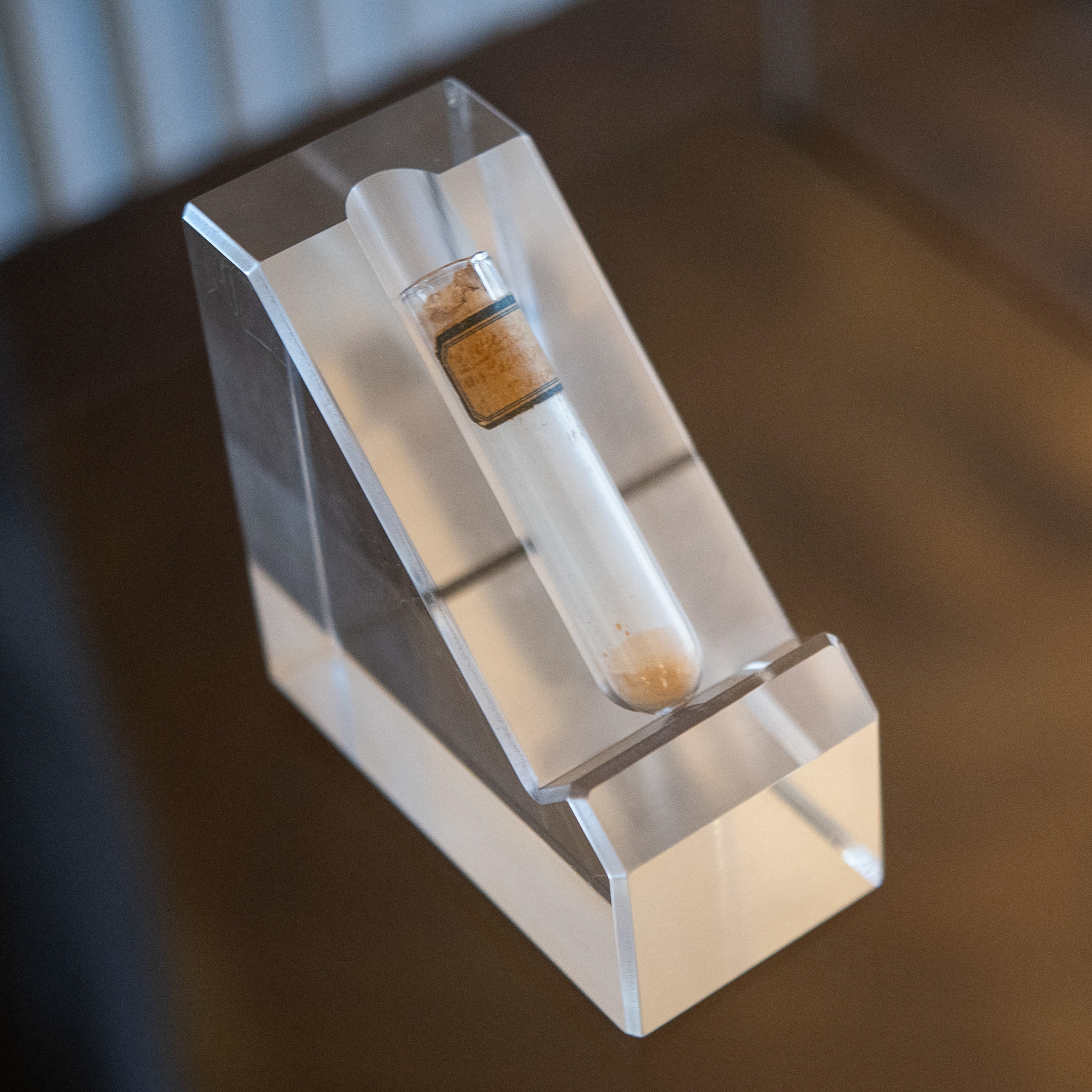 A clear acrylic stand holds a small glass vial with a cork stopper, containing a light brown, crumbly substance.