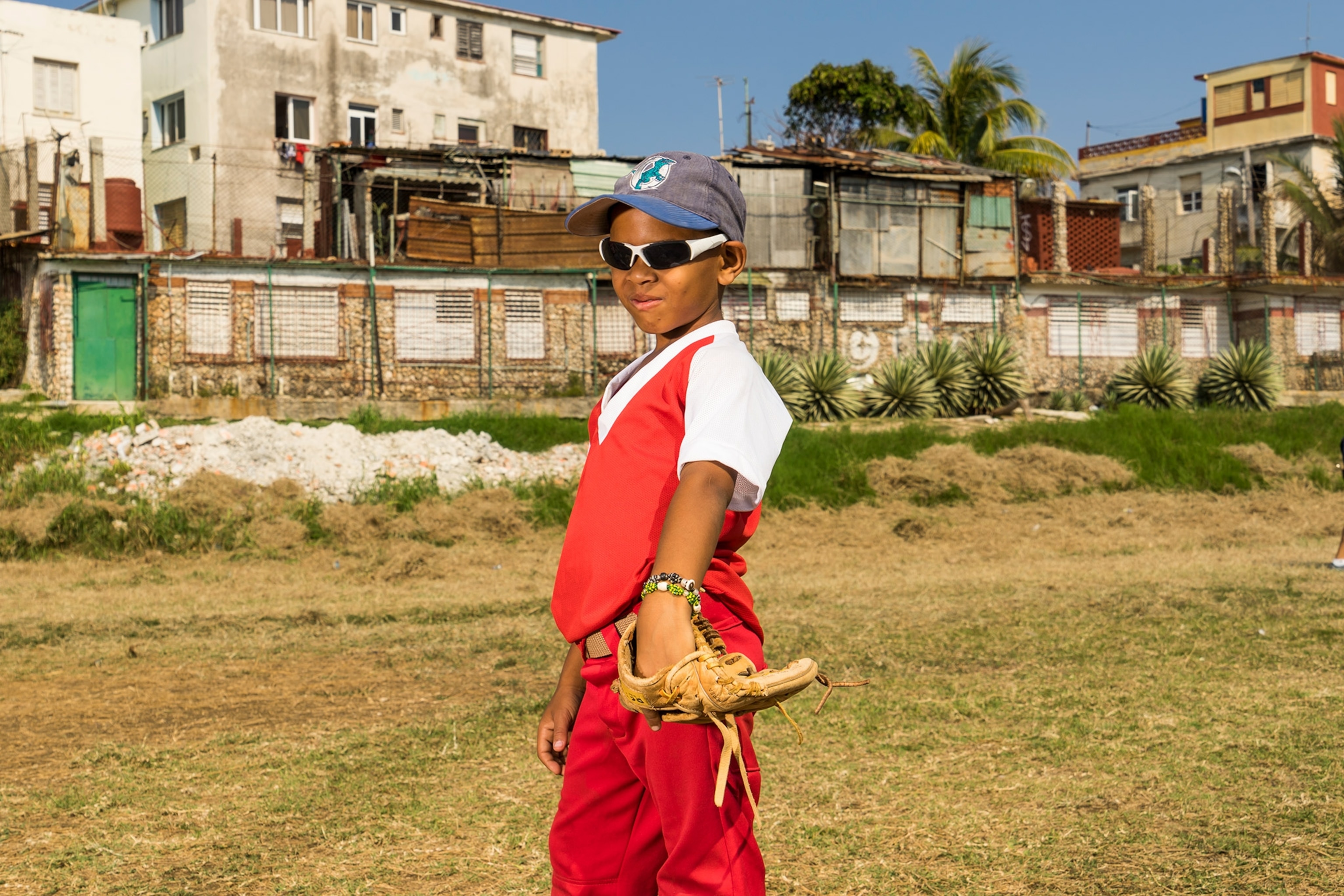 A boy wears a red and white baseball uniform with a blue had and poses with white shades.