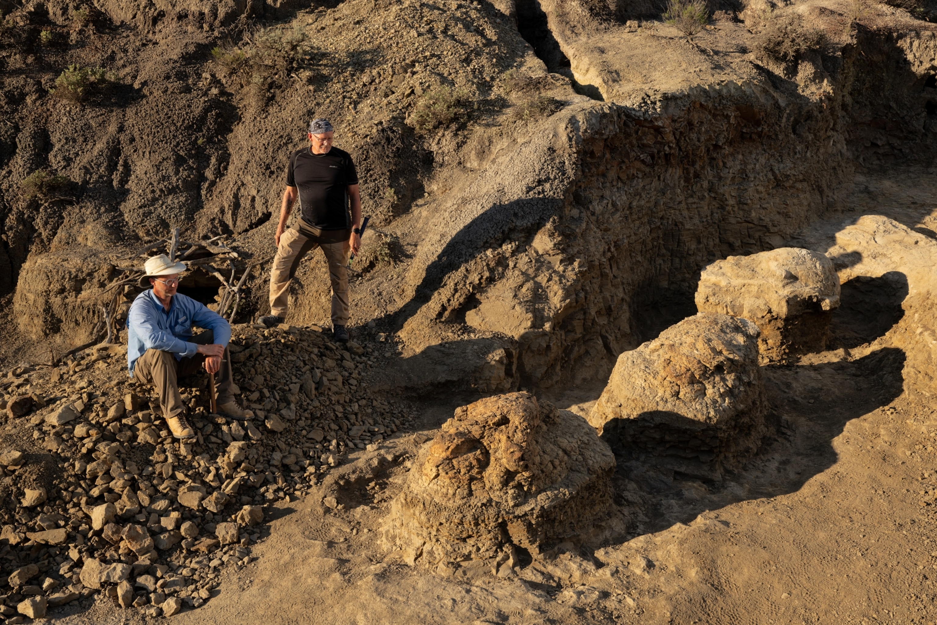 Two men overlook a sunlit, rugged fossil formation; one sits on dark scree while the other stands nearby, creating an adventurous mood.