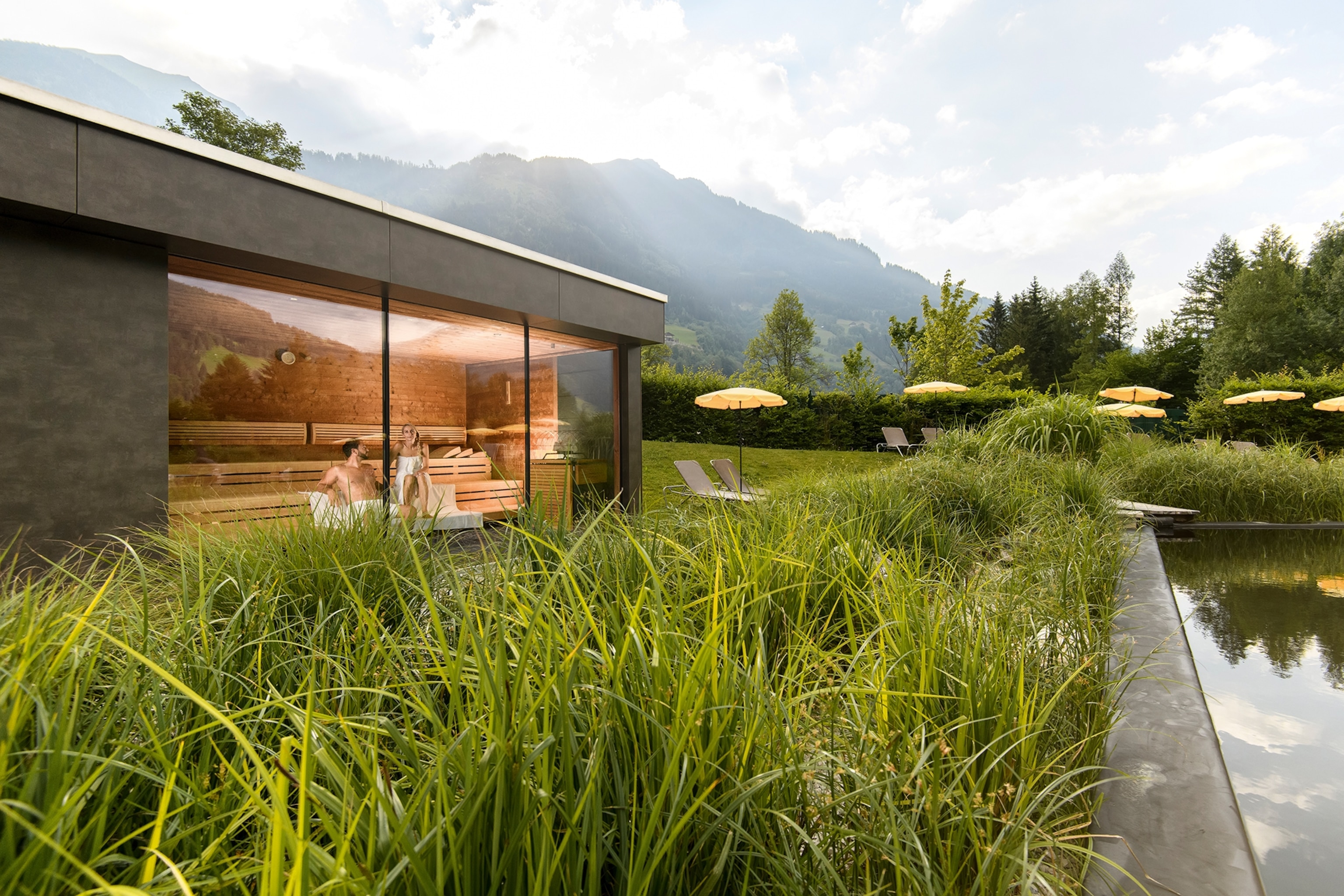 A couple lounging in a glass-windowed sauna in the middle of a mountain valley with a pool on the side.