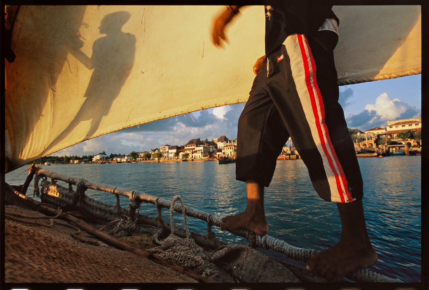 Picture of person on sailboat and the shadow on another person on the sail. The port town on the background.