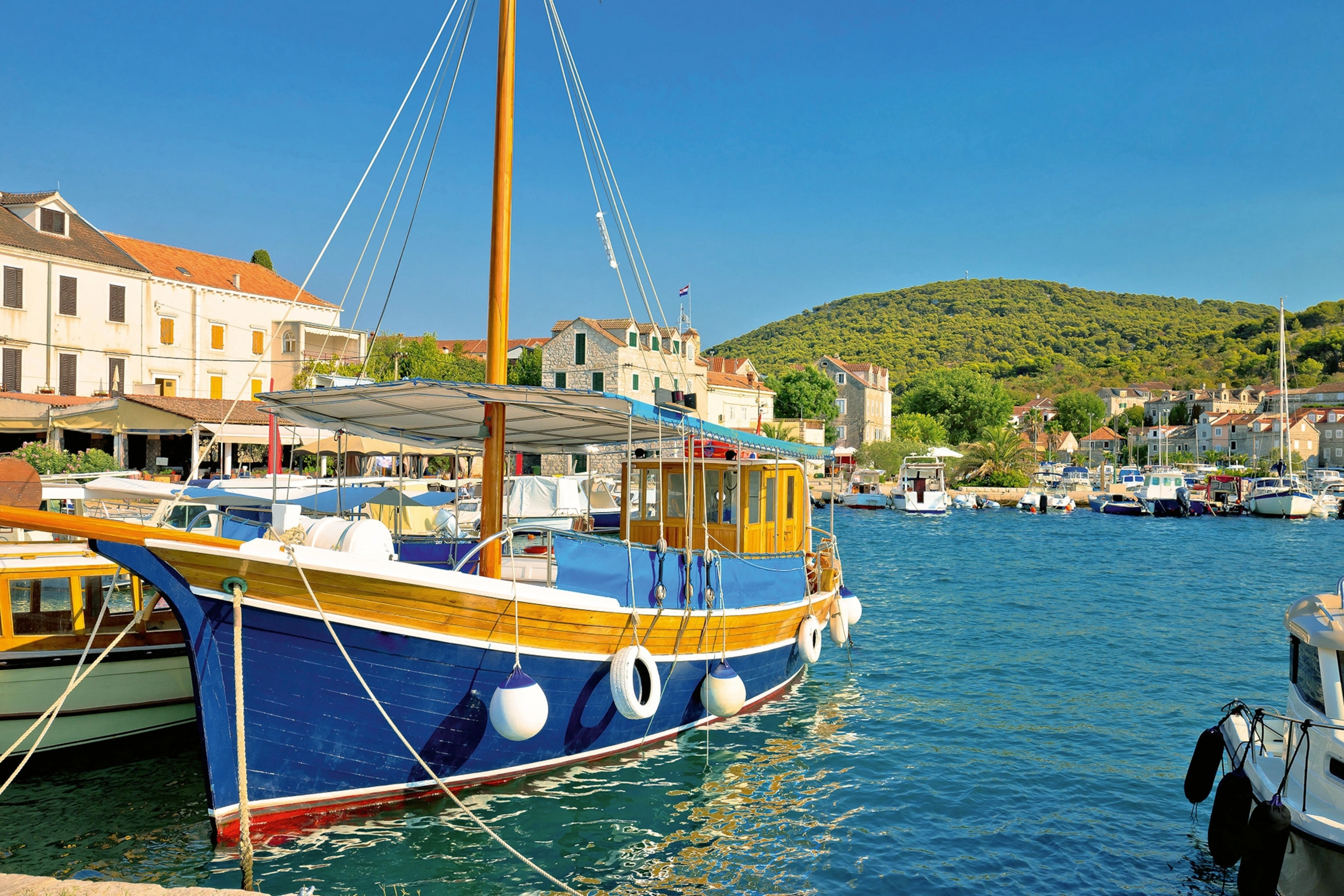 A wooden, sailing ferry anchored in the harbour of a medieval stone town with the ocean in the background.