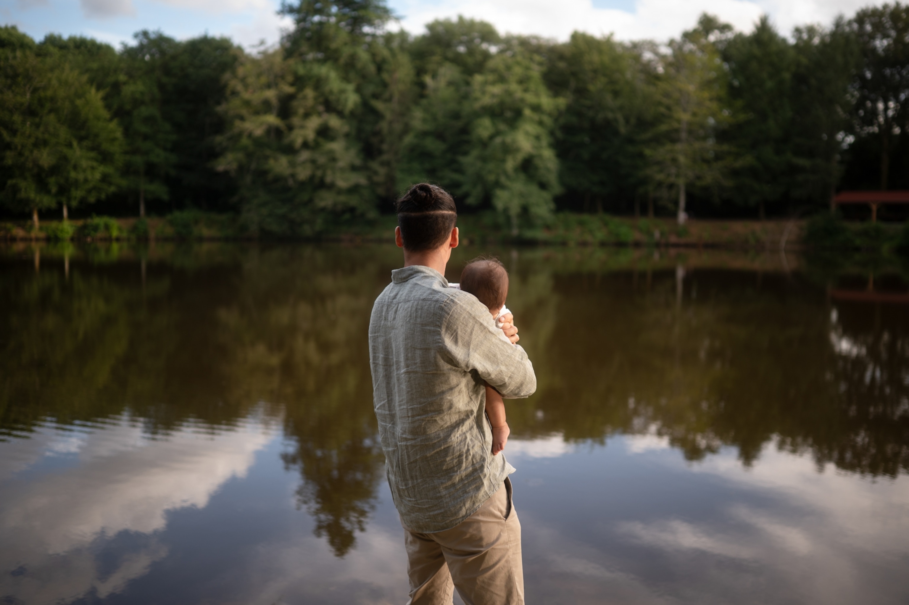 A man stands by a calm lake holding a baby, facing lush green trees reflected in the water