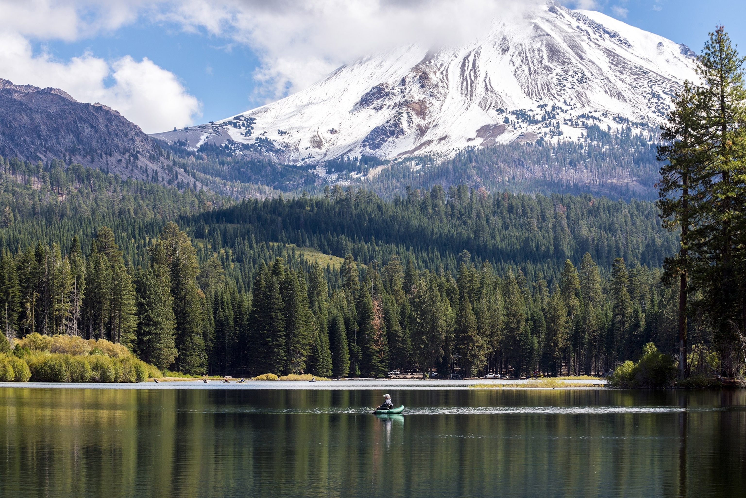 A boat floats in the middle of a lake, surrounded by woods, a large snow-covered mountain towers behind.